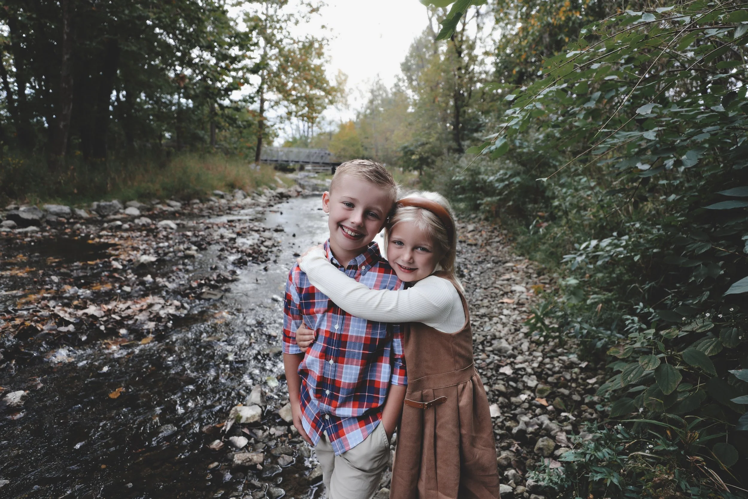 Two children, a boy and a girl, hugging each other near a creek in a wooded area.
