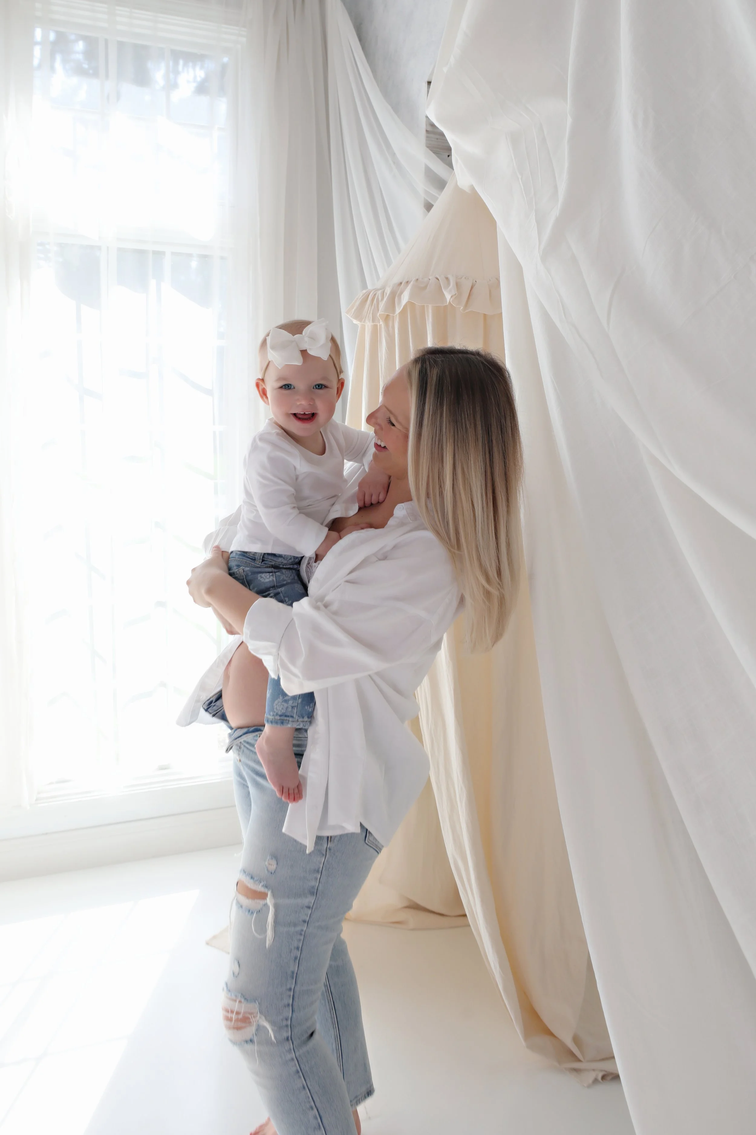 A woman with blonde hair is holding a smiling toddler girl with a big white bow in her hair, in a bright room with sheer curtains and a canopy.