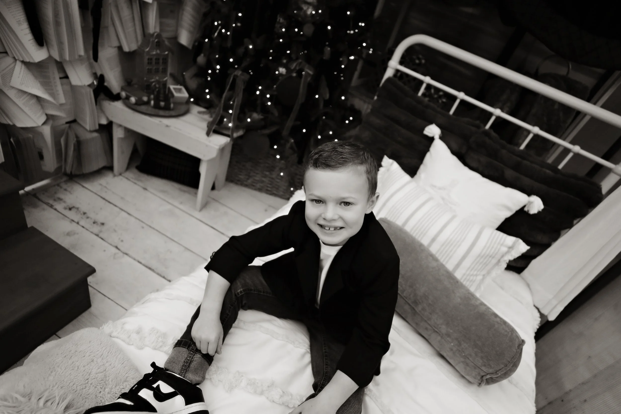A young boy sitting on a bed, smiling at the camera, in a cozy, decorated room with pillows, wooden furniture, and Christmas lights in the background.