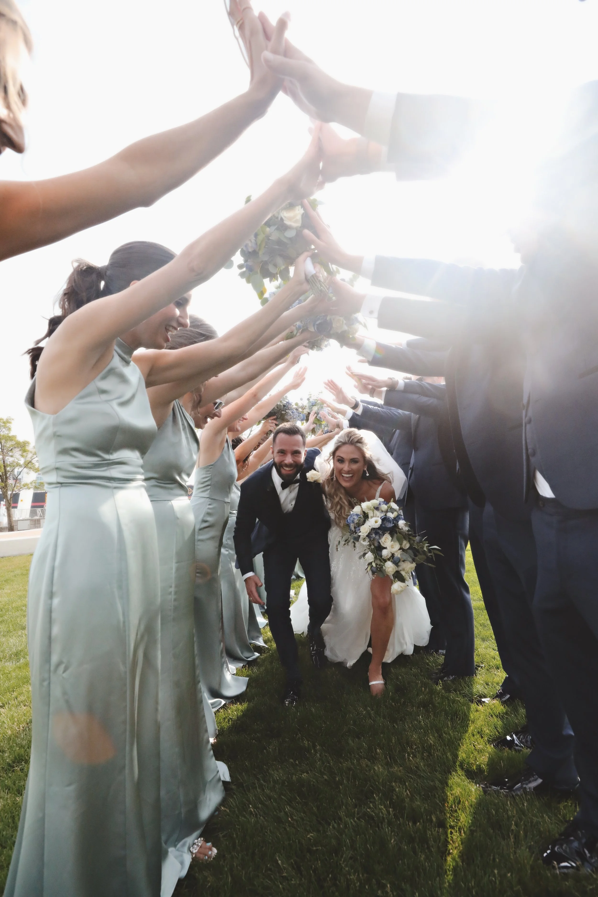 Bride and groom smiling as they walk through an arch of raised hands and bouquets during their wedding celebration outdoors.