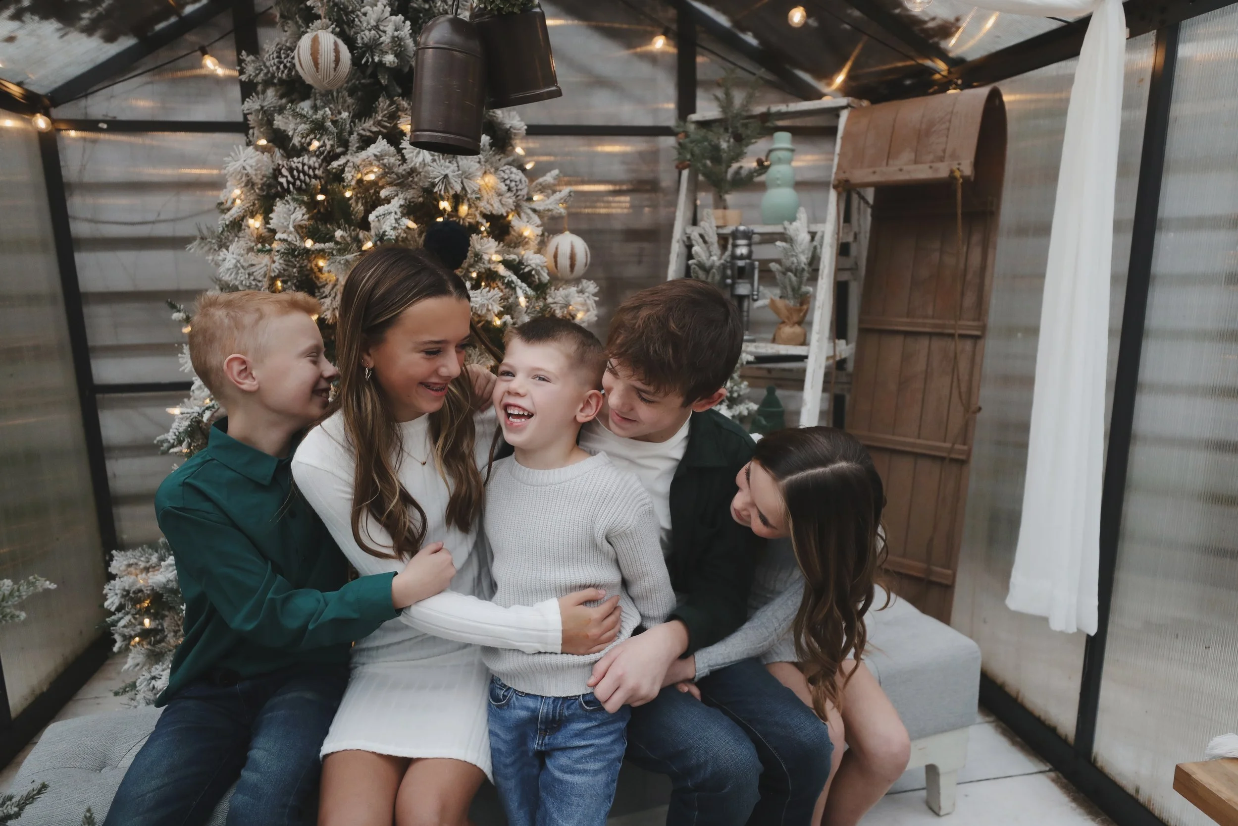 A group of five children and teenagers sitting together in front of a Christmas tree with ornaments and lights, smiling and laughing.