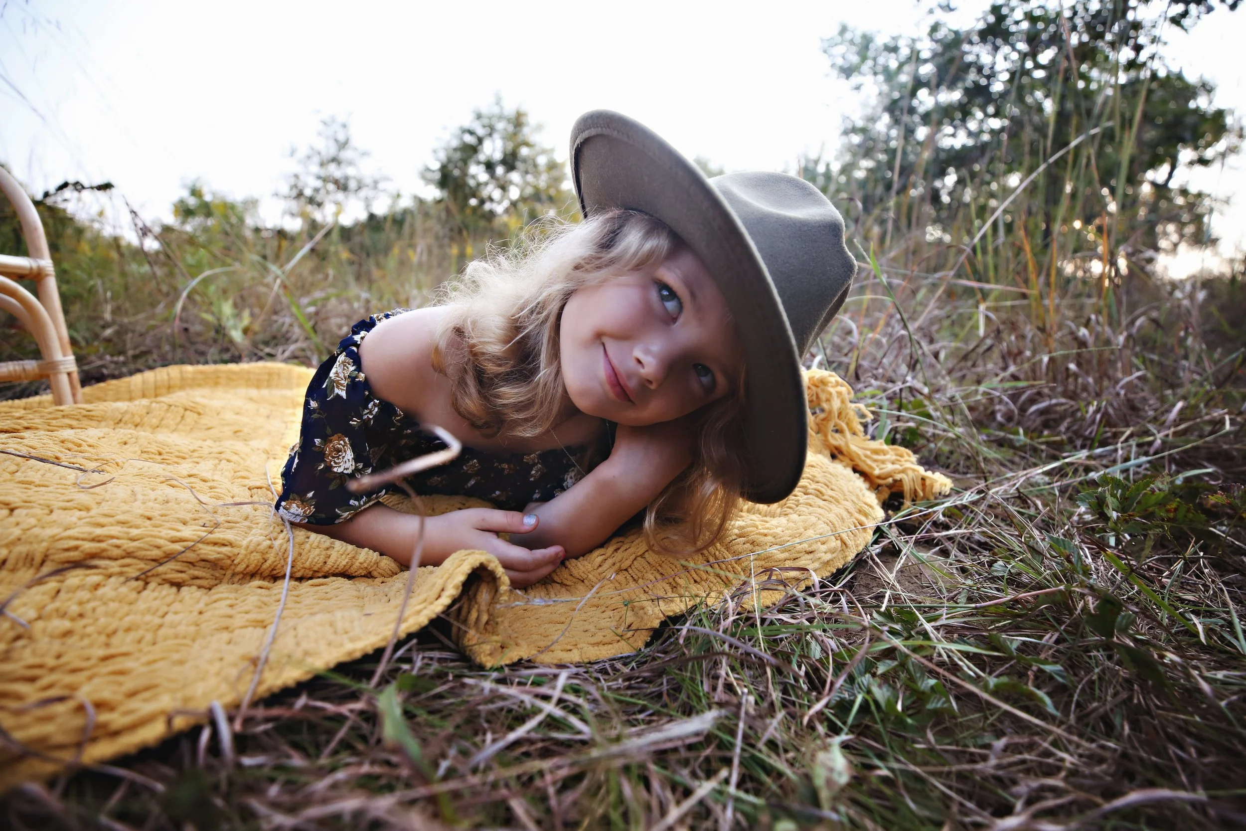 A young girl with blonde hair, wearing a gray hat and a floral dress, lying on a yellow blanket outdoors in a grassy area with trees in the background, resting her head on her arm and looking at the camera.