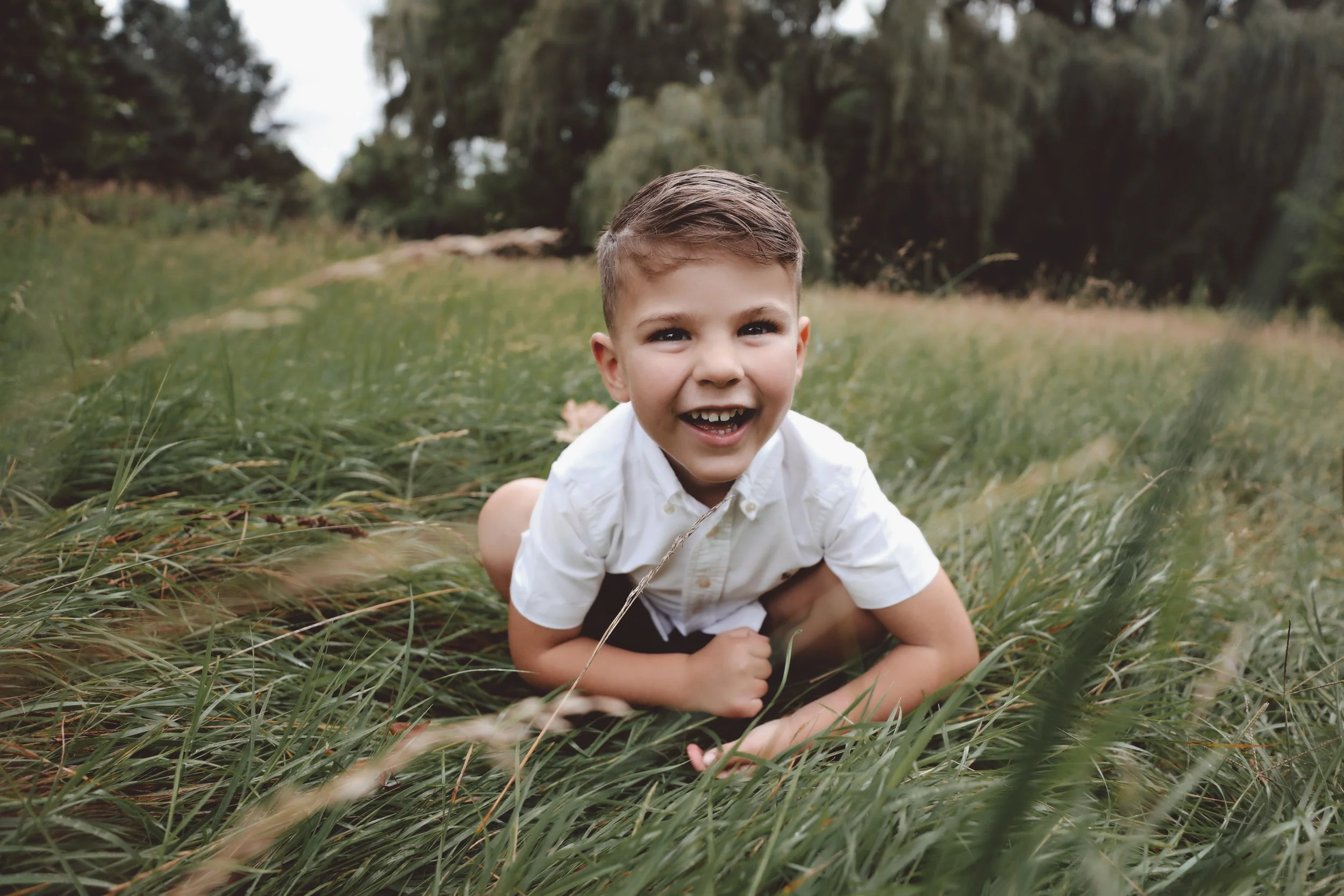 A young boy with short brown hair and a white shirt crawling on the grass in a field, smiling at the camera.