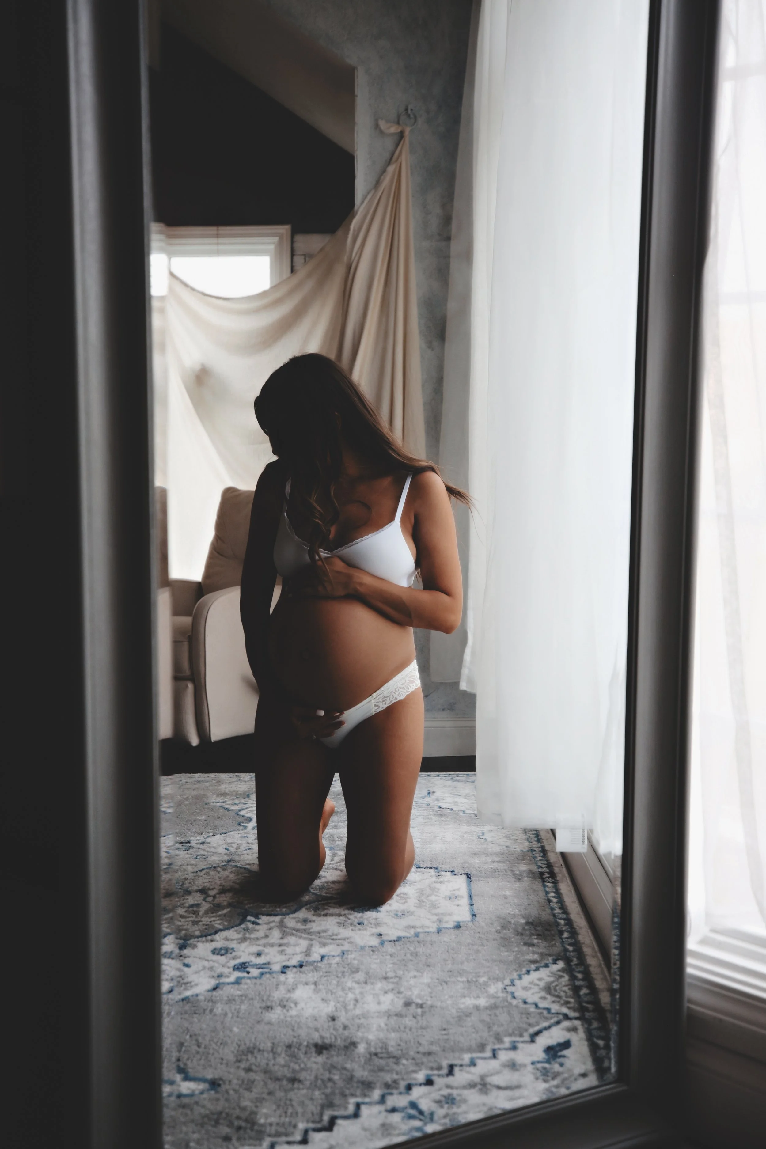 A pregnant woman kneeling on a rug in front of a mirror, wearing white lingerie, gently touching her belly, with natural light coming through sheer curtains.