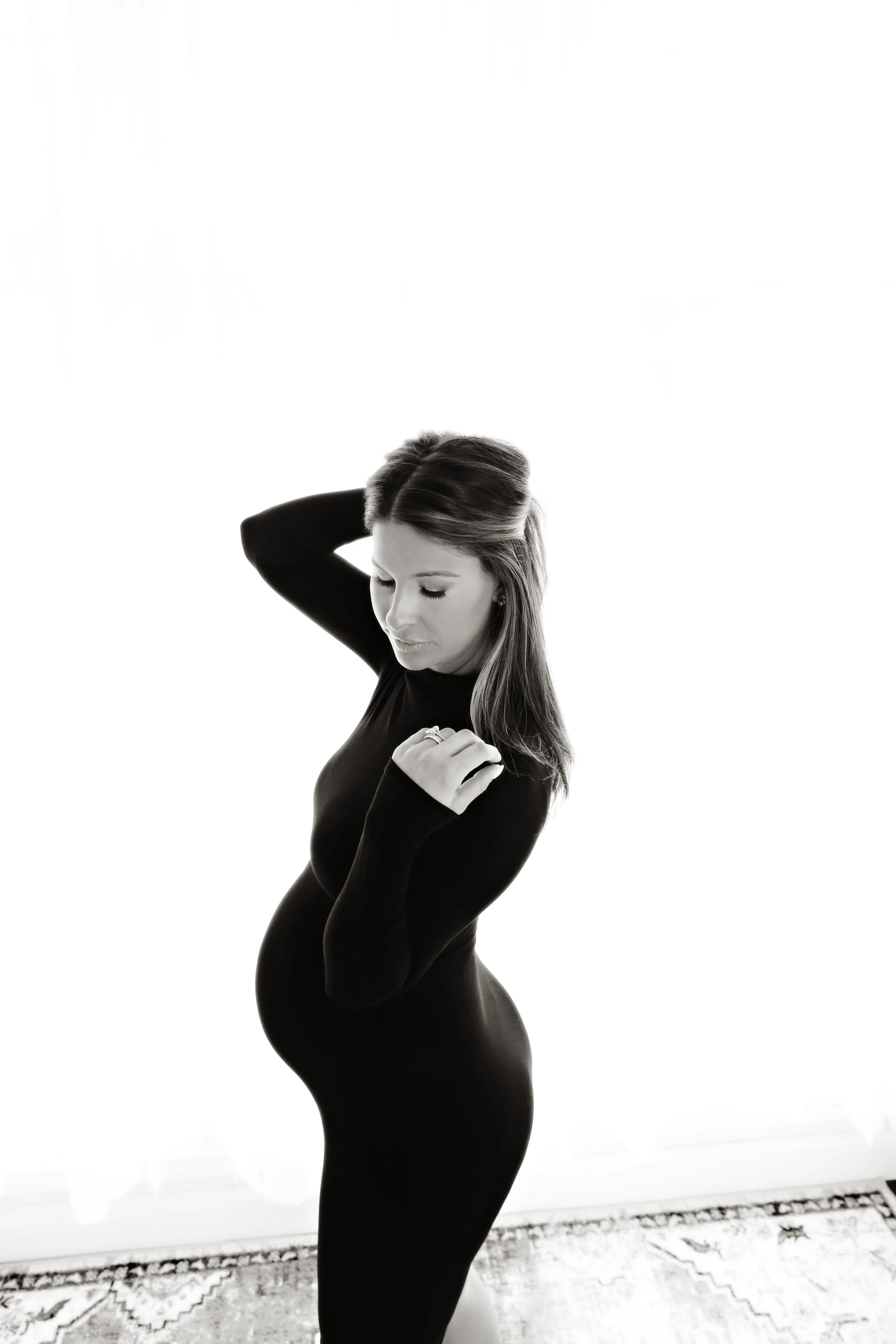 Black and white photo of woman in a fitted, long-sleeve dress with long hair, standing indoors near a window with patterned rug at her feet, in a reflective or contemplative pose.