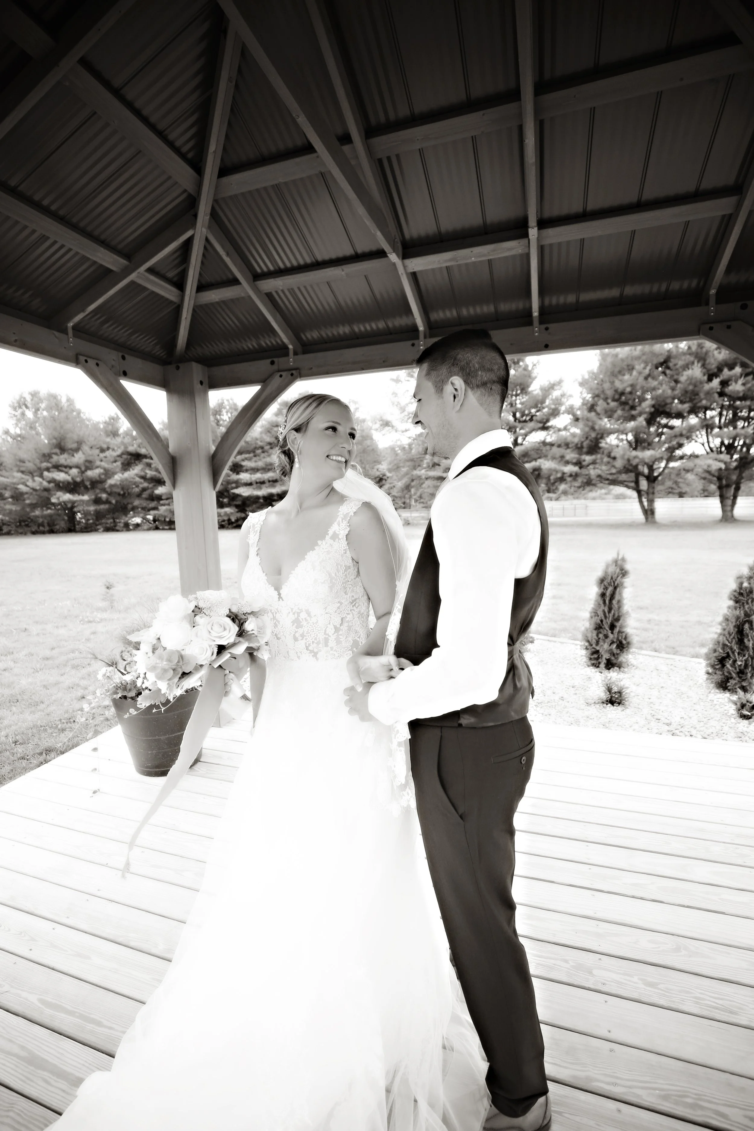 Black and white photo of a bride and groom happily looking at each other under a gazebo on their wedding day. The bride is holding a bouquet and wearing a lace wedding gown. The groom is dressed in a vest and dress shirt. There are trees and small bu