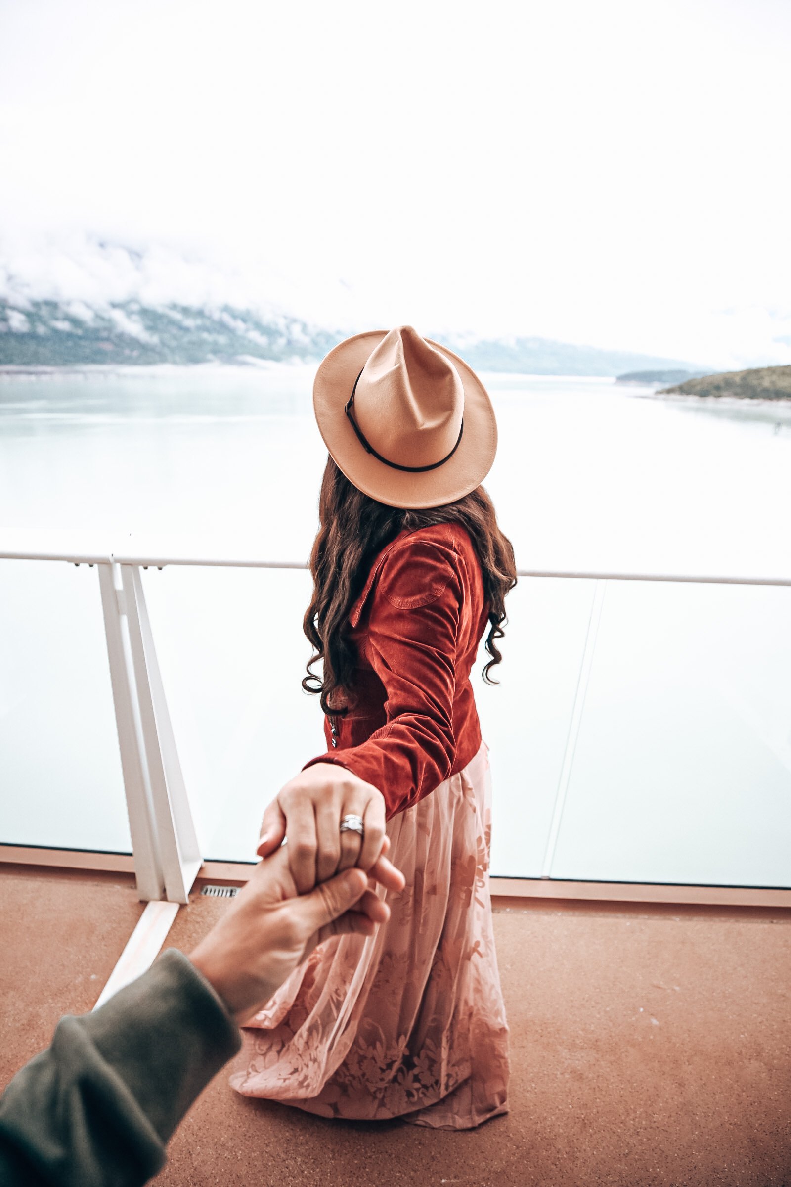 A woman with long, wavy hair wearing a wide-brimmed tan hat, a red jacket, and a long, patterned pink skirt, holding hands with the viewer on a boat with a mountain and lake in the background.
