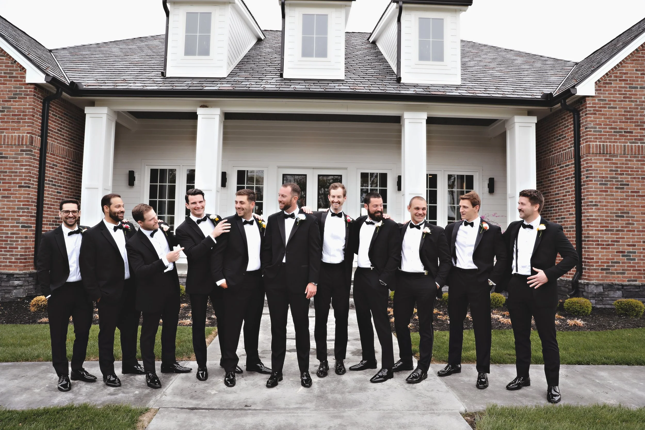 A group of eleven men in tuxedos standing outside in front of a house, smiling and socializing.