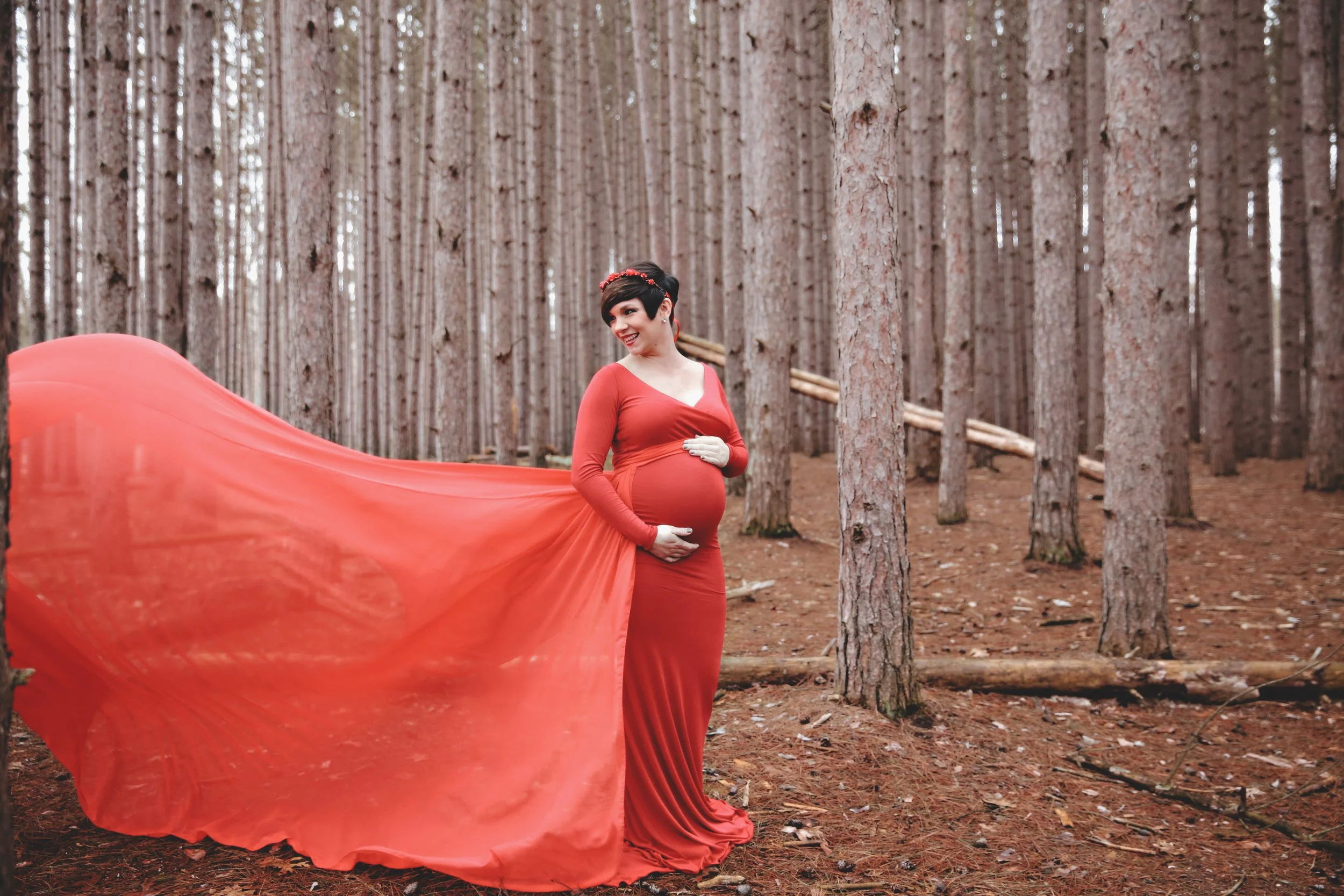 A pregnant woman in a long red dress with a flowing train in a forest of tall, straight pine trees, smiling and gently holding her belly.