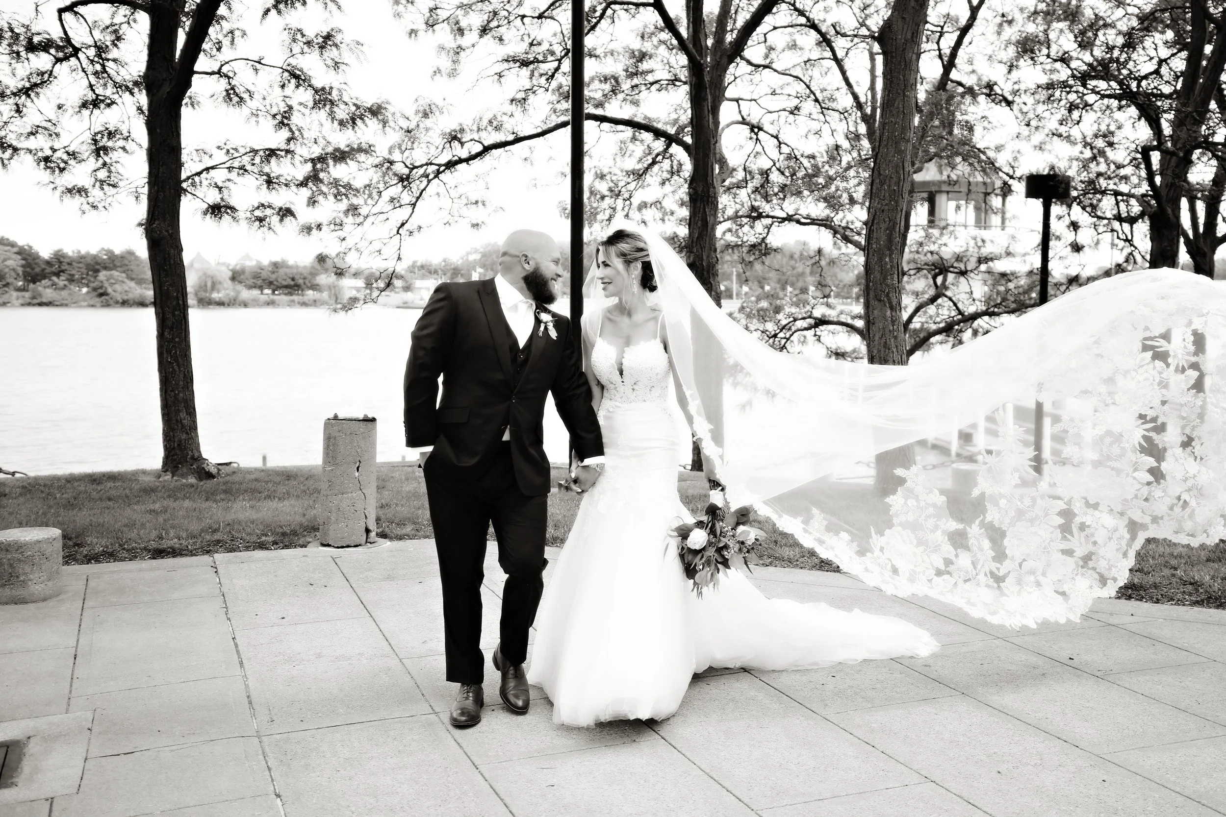 Black and white photo of a bride and groom holding hands and walking outdoors near a body of water, with trees in the background.