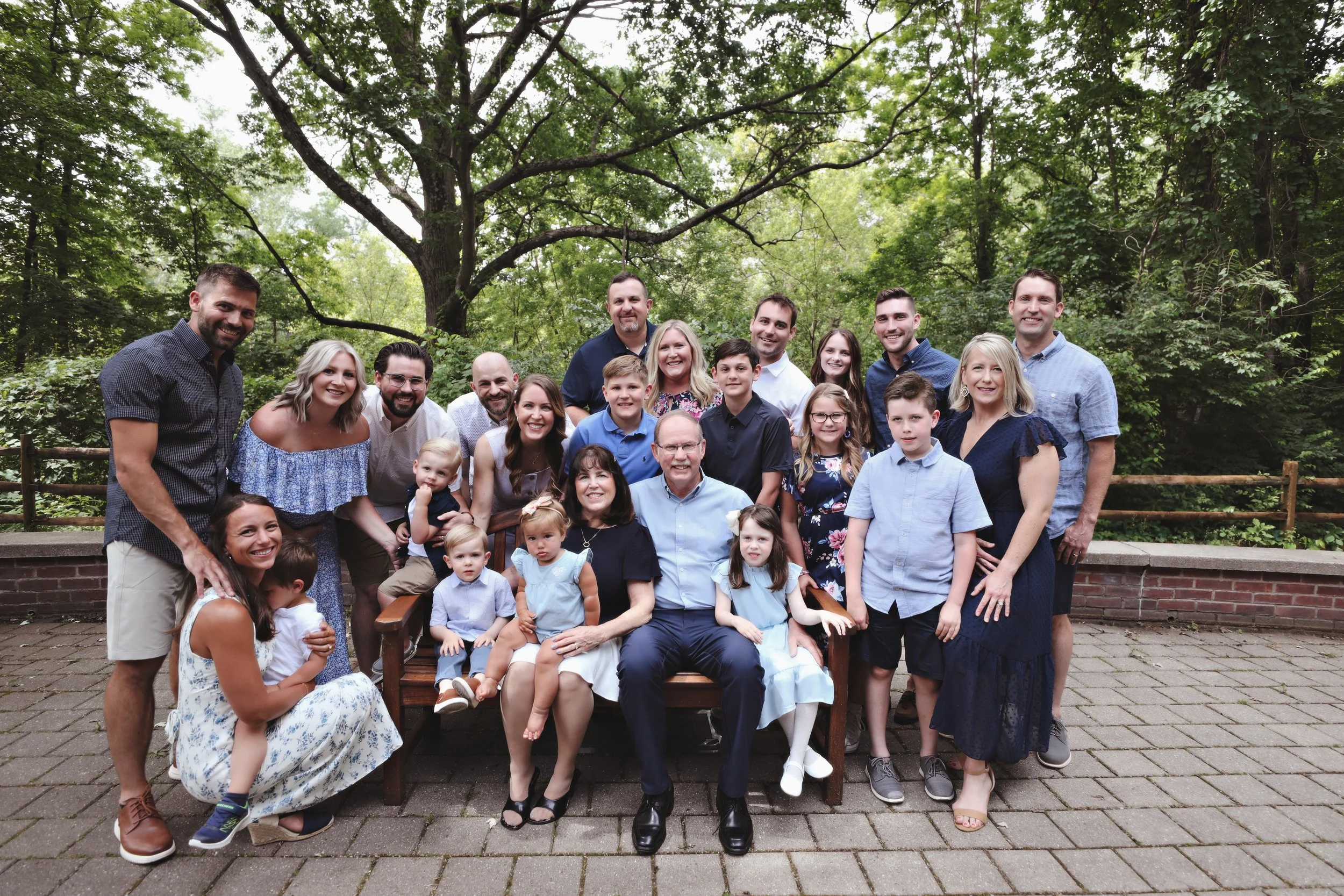 Family group photo outdoors with adults and children, some sitting and some standing, on a paved area surrounded by trees.