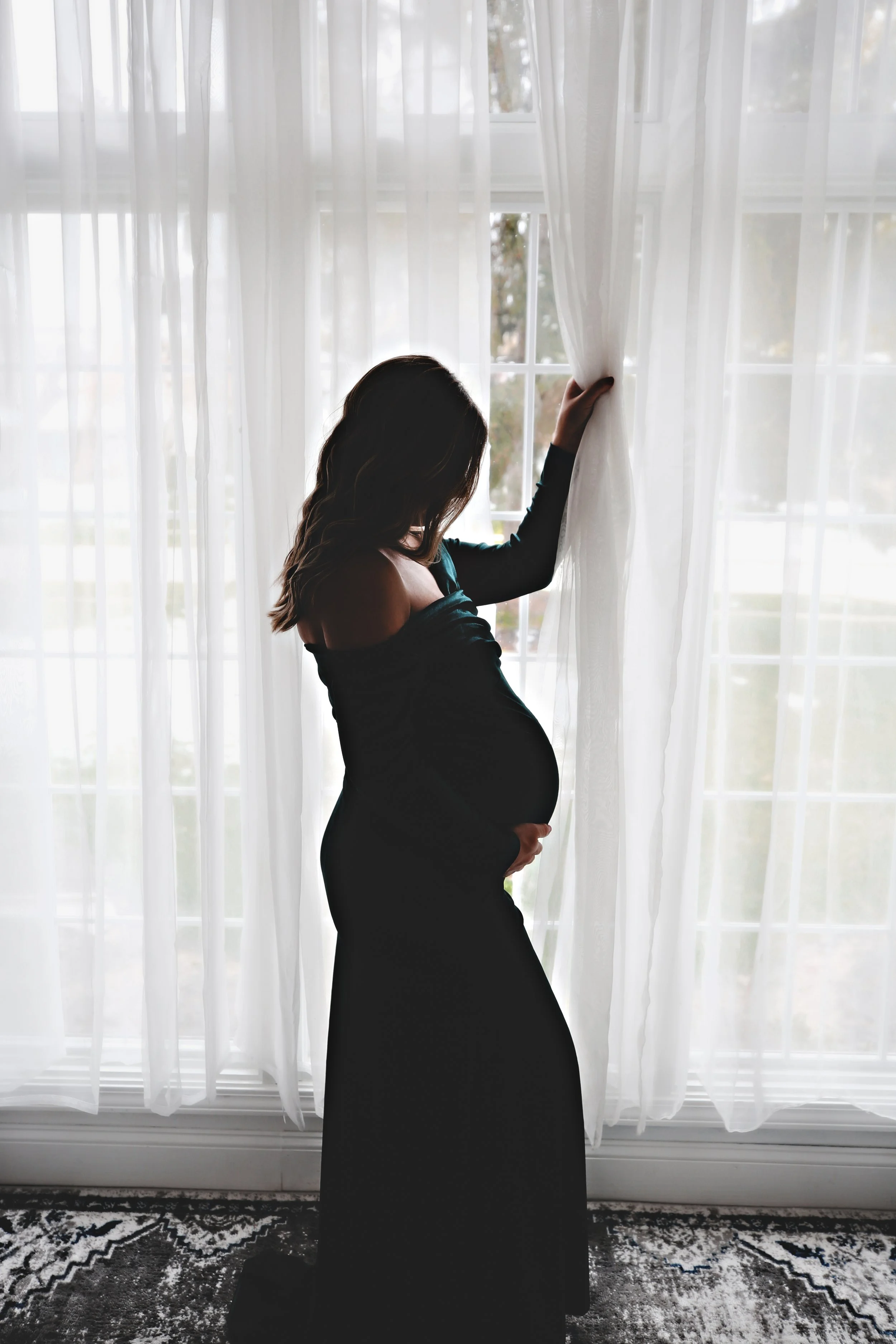 Silhouette of a pregnant woman in a black dress standing by window, holding curtain with one hand and resting her other hand on her belly.