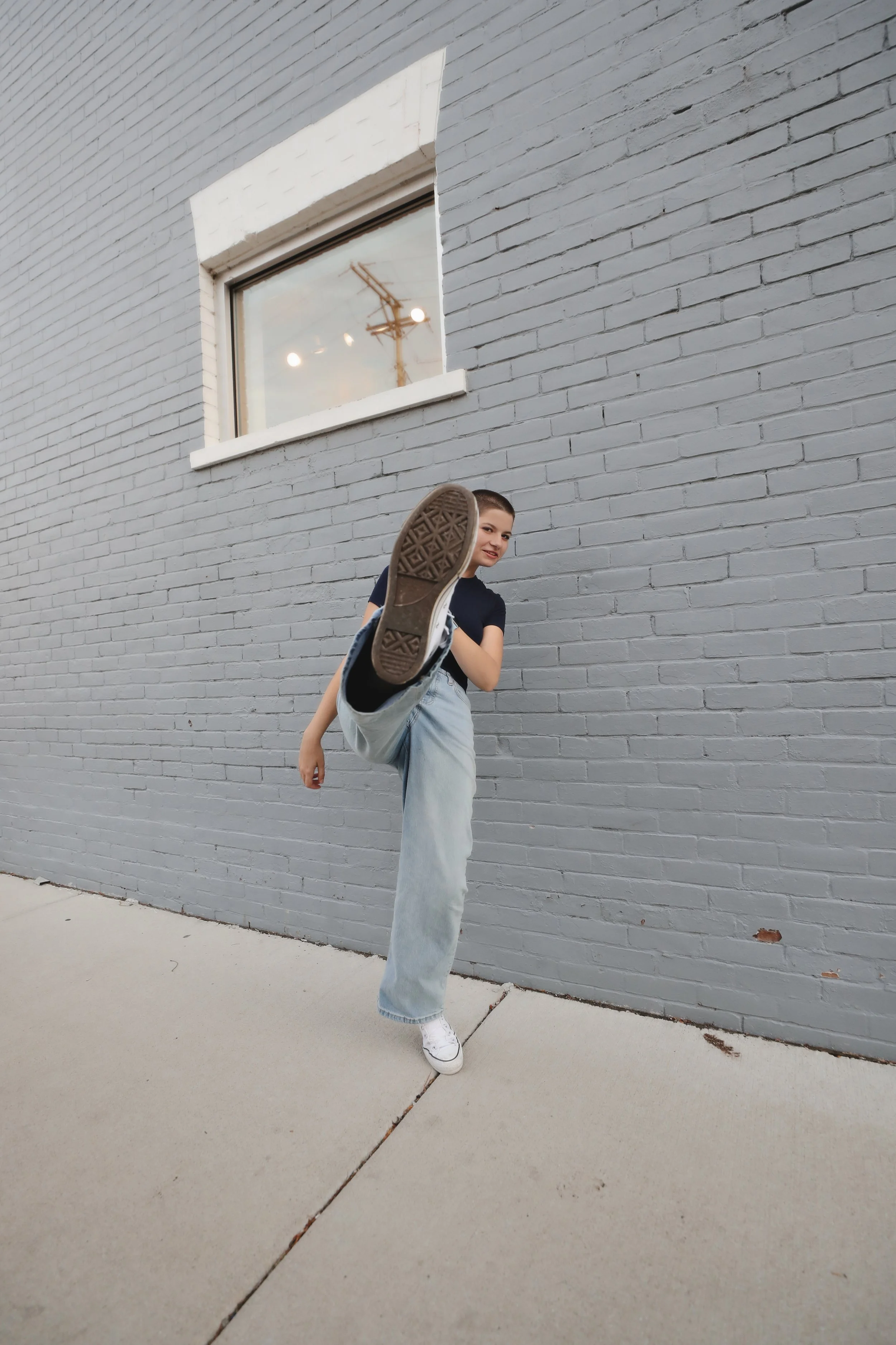 A young woman standing outdoors against a gray brick wall, dressed in a black t-shirt, light-washed jeans, and white sneakers, performing a high kick with her left leg extended towards the camera.