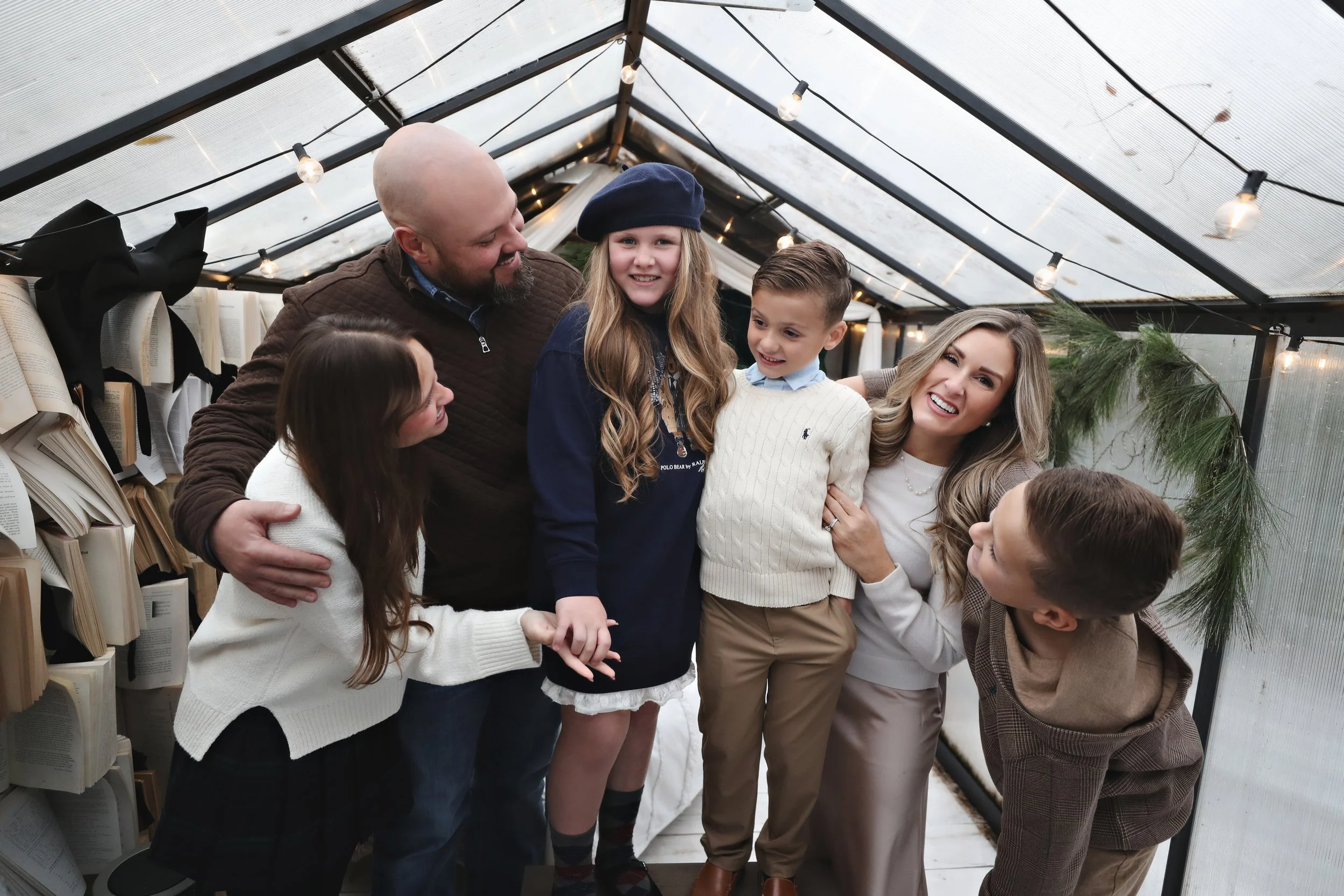 A group of six people, including three children and three adults, smiling and embracing in a greenhouse decorated with string lights and books.