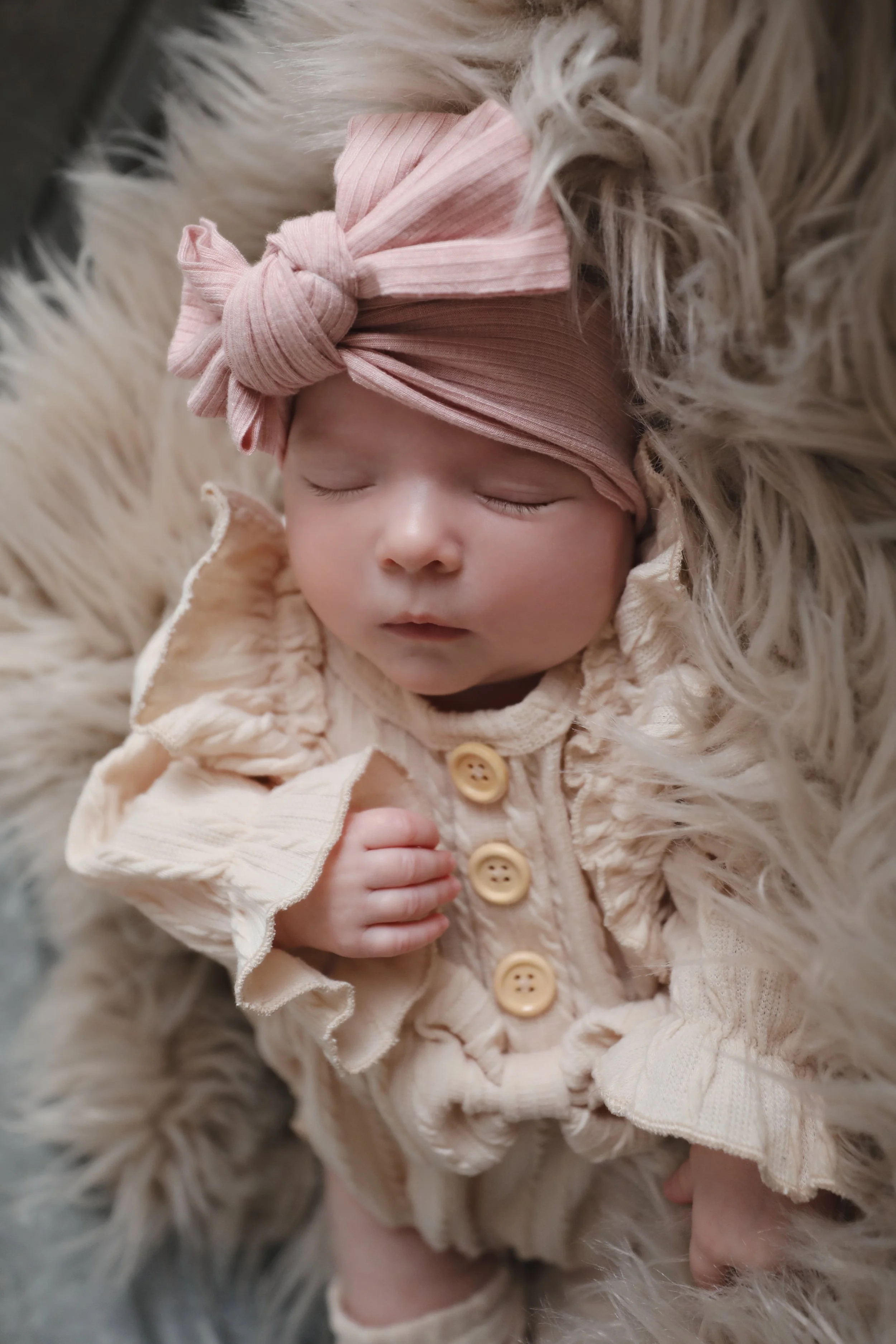 Close-up of a sleeping baby girl wearing a pink headband with a bow, dressed in a cream-colored outfit with buttons, lying on a fluffy faux fur surface.