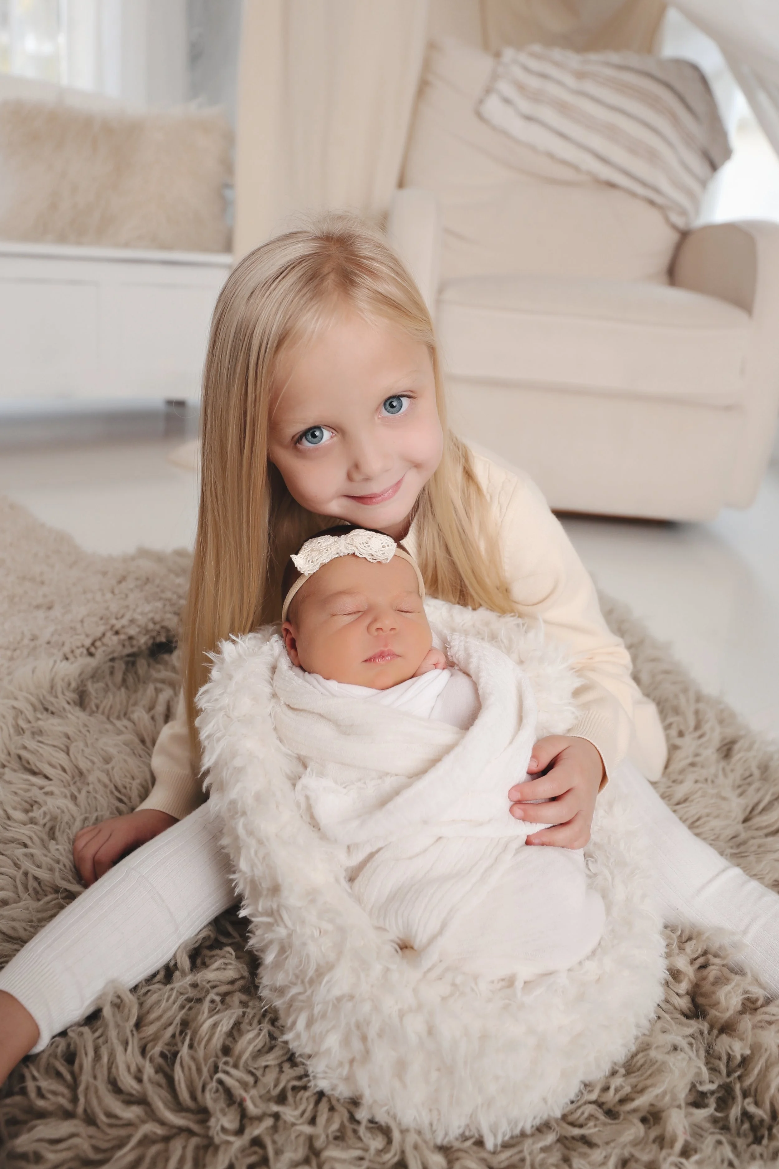 A young girl with long blonde hair and blue eyes holding a newborn baby wrapped in a white blanket inside a cozy, cream-colored room.