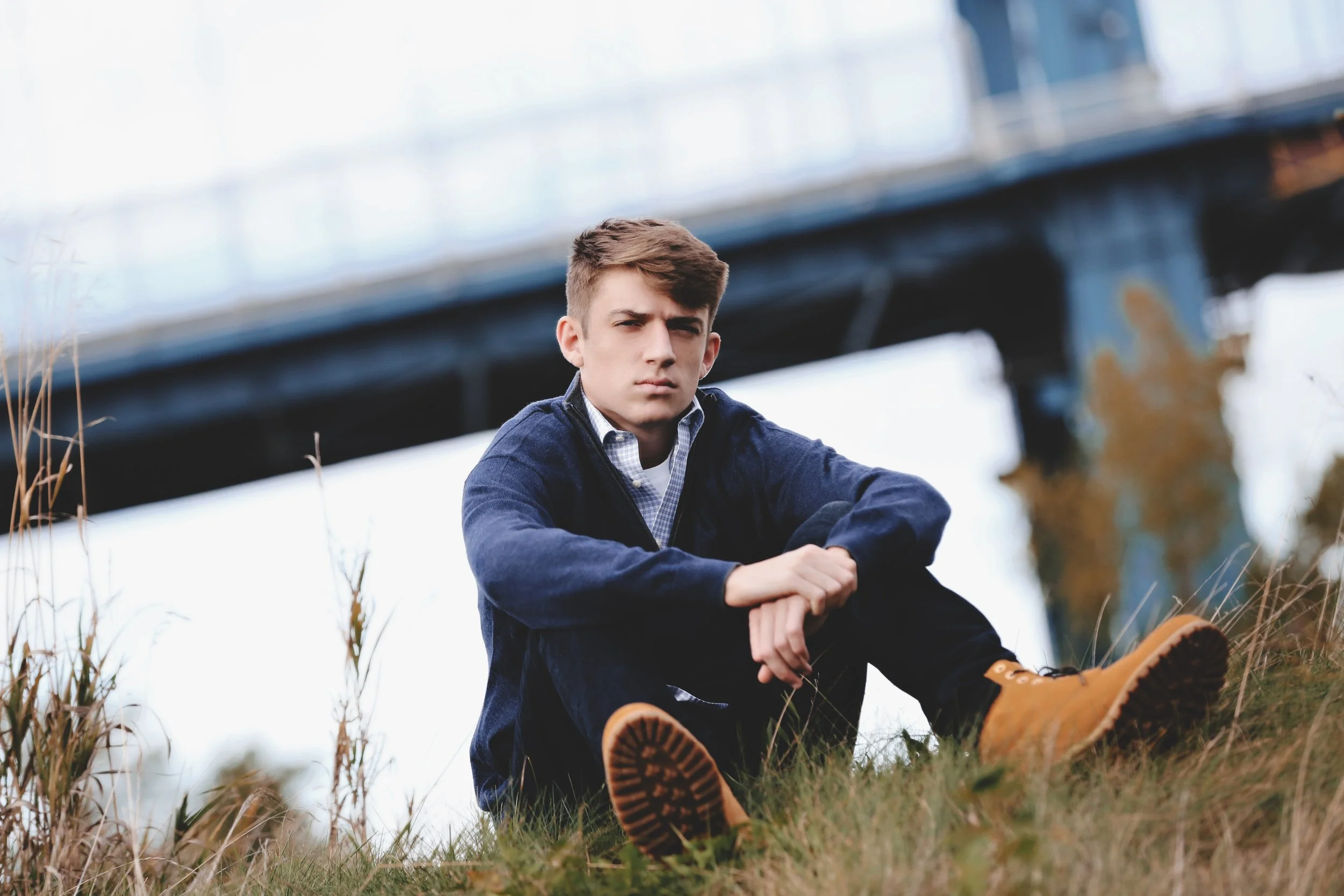 A young man with brown hair sitting outdoors on grass, wearing a navy jacket, checkered shirt, black pants, and tan boots, with a bridge in the background.