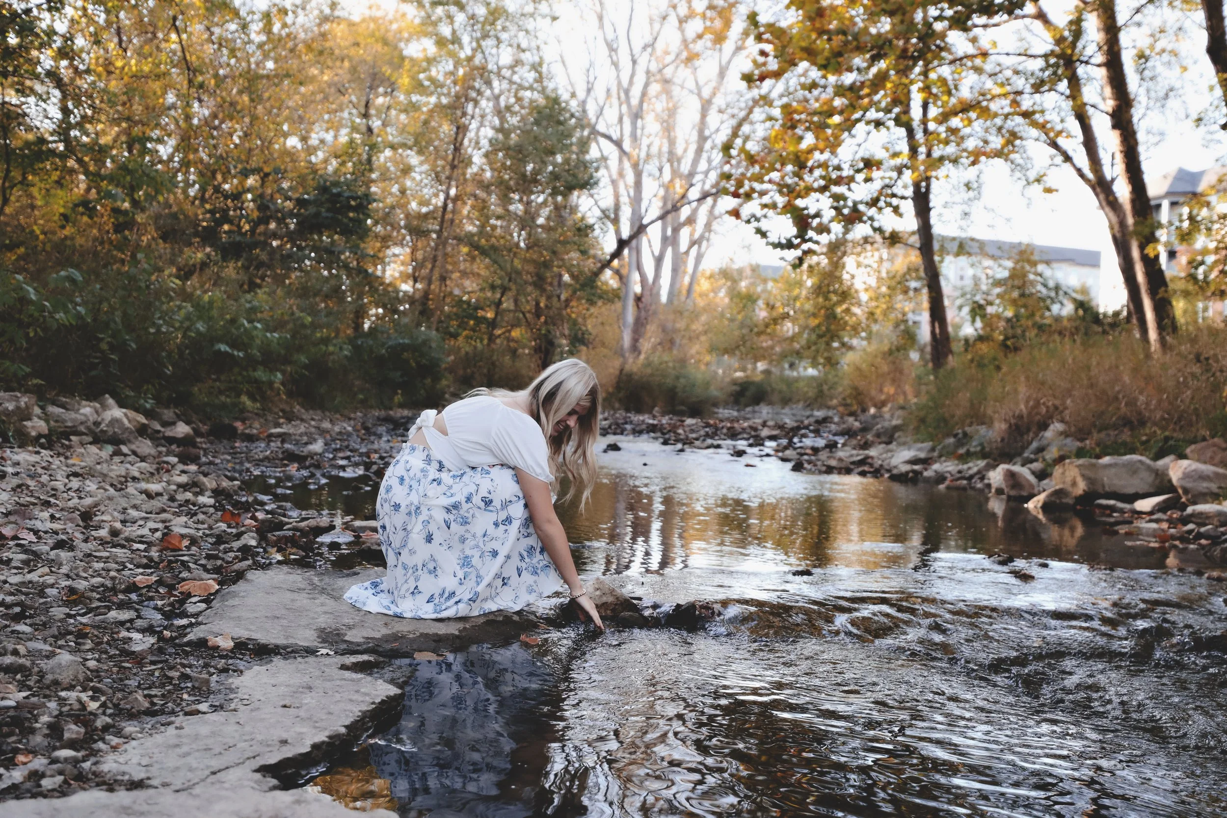 A woman with long blonde hair wearing a white top and a white skirt with blue floral patterns, kneeling by a small creek in a wooded area during autumn, touching the water.