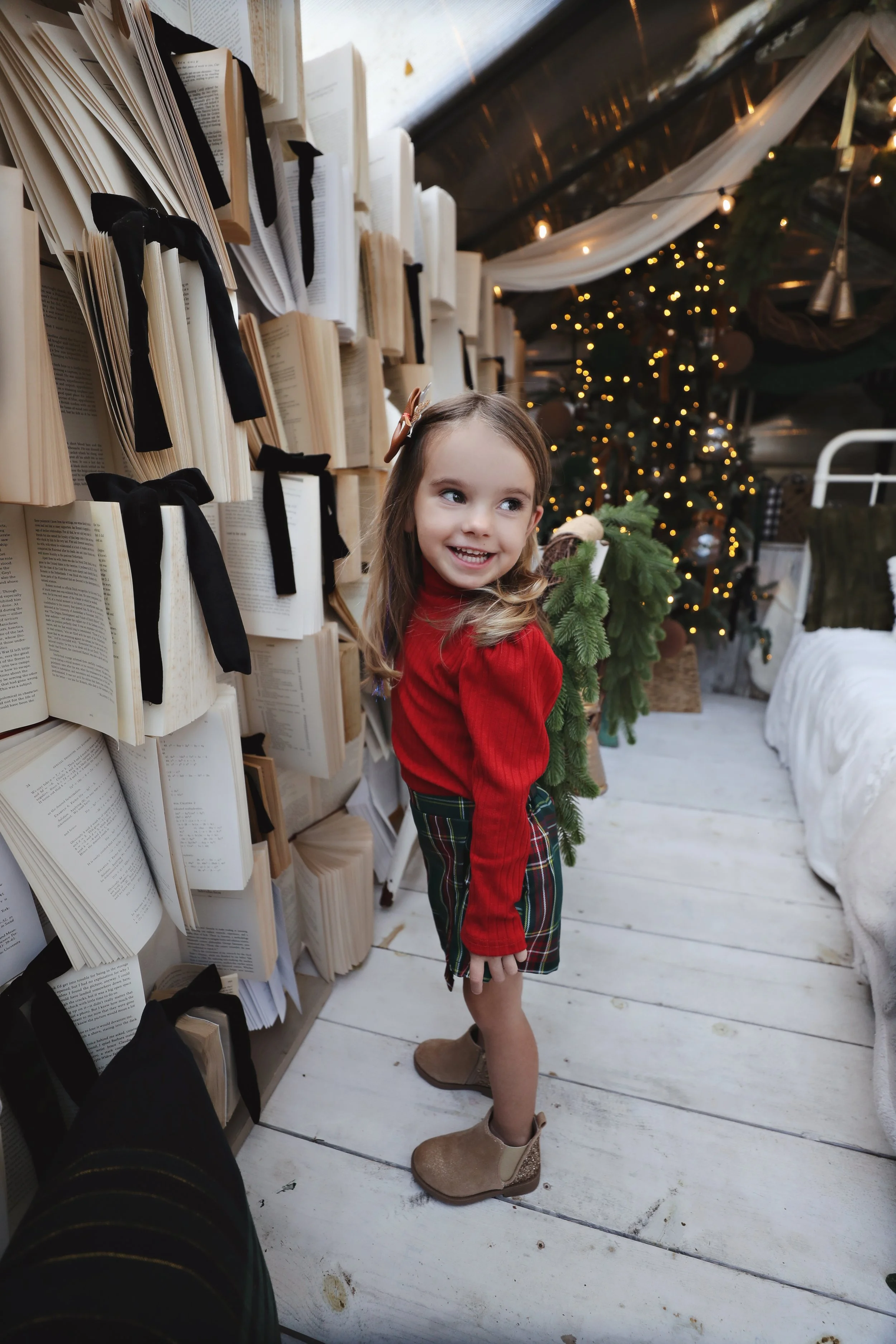 A young girl in a red sweater and plaid skirt standing next to a bookshelf filled with open books tied with black ribbons, smiling at the camera. A Christmas tree decorated with lights and ornaments is visible in the background.