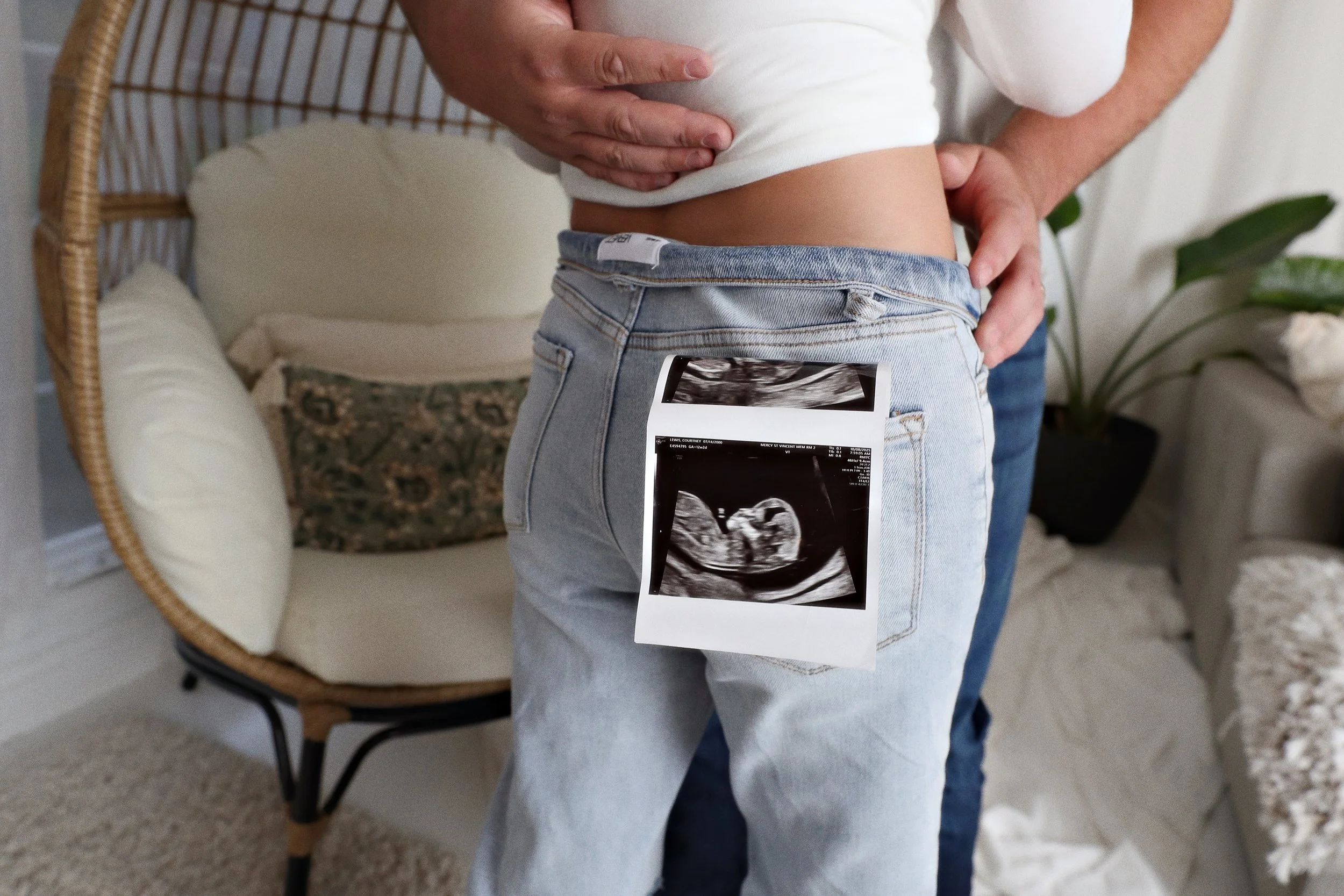Person holding ultrasound picture of a fetus in front of their abdomen, with a chair, pillow, and houseplant in the background.
