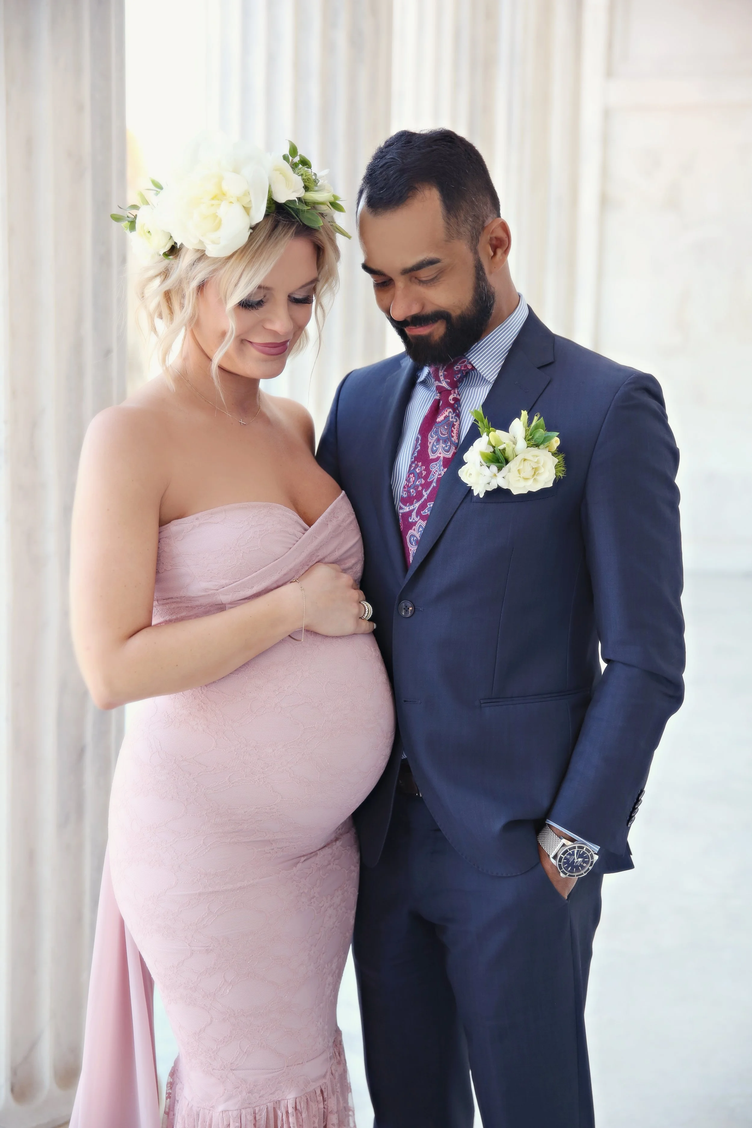 A pregnant woman in a pink strapless dress and a flower crown stands with a man in a dark blue suit, both smiling and looking down at her belly, in a bright indoor setting.