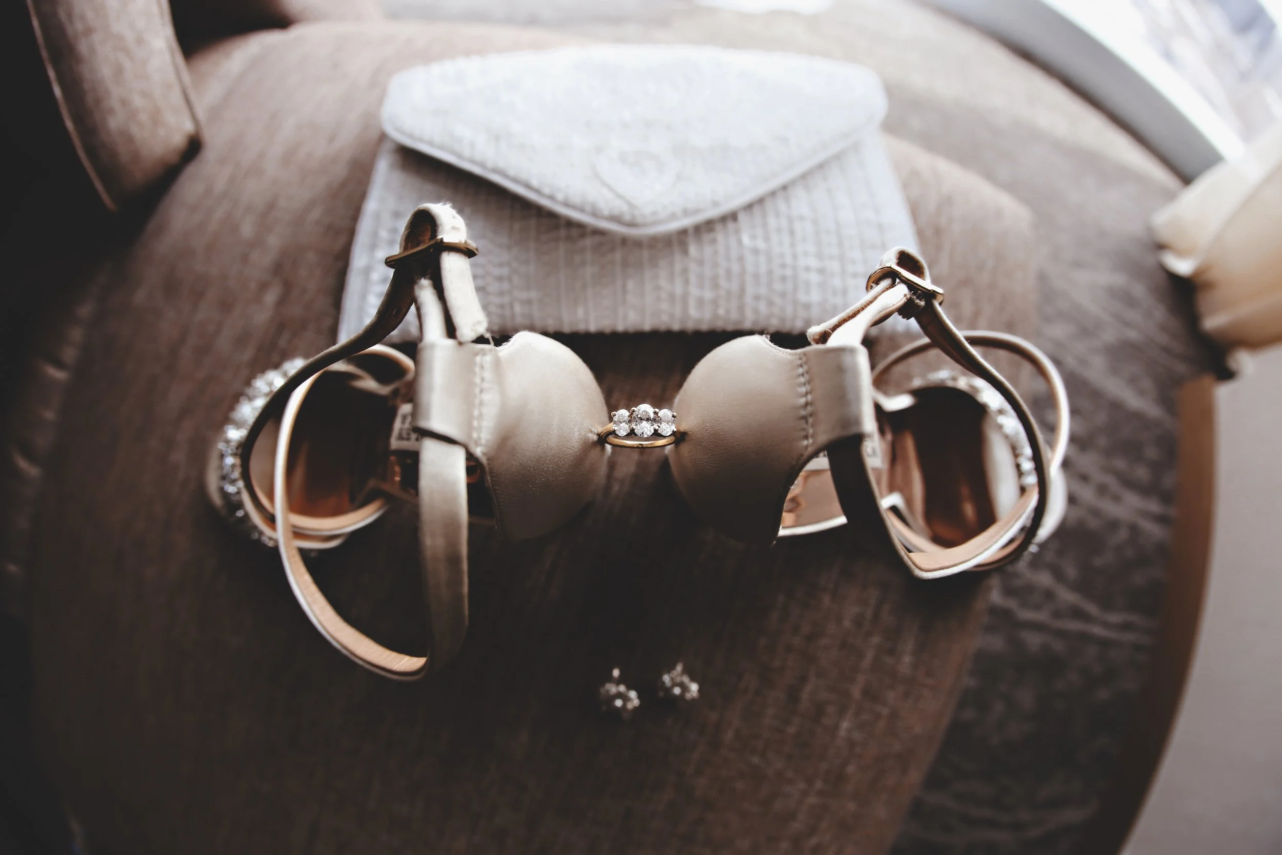 A pair of beige high-heeled shoes with ankle straps, a ring with three sparkly stones, and matching earrings on a brown fabric surface.