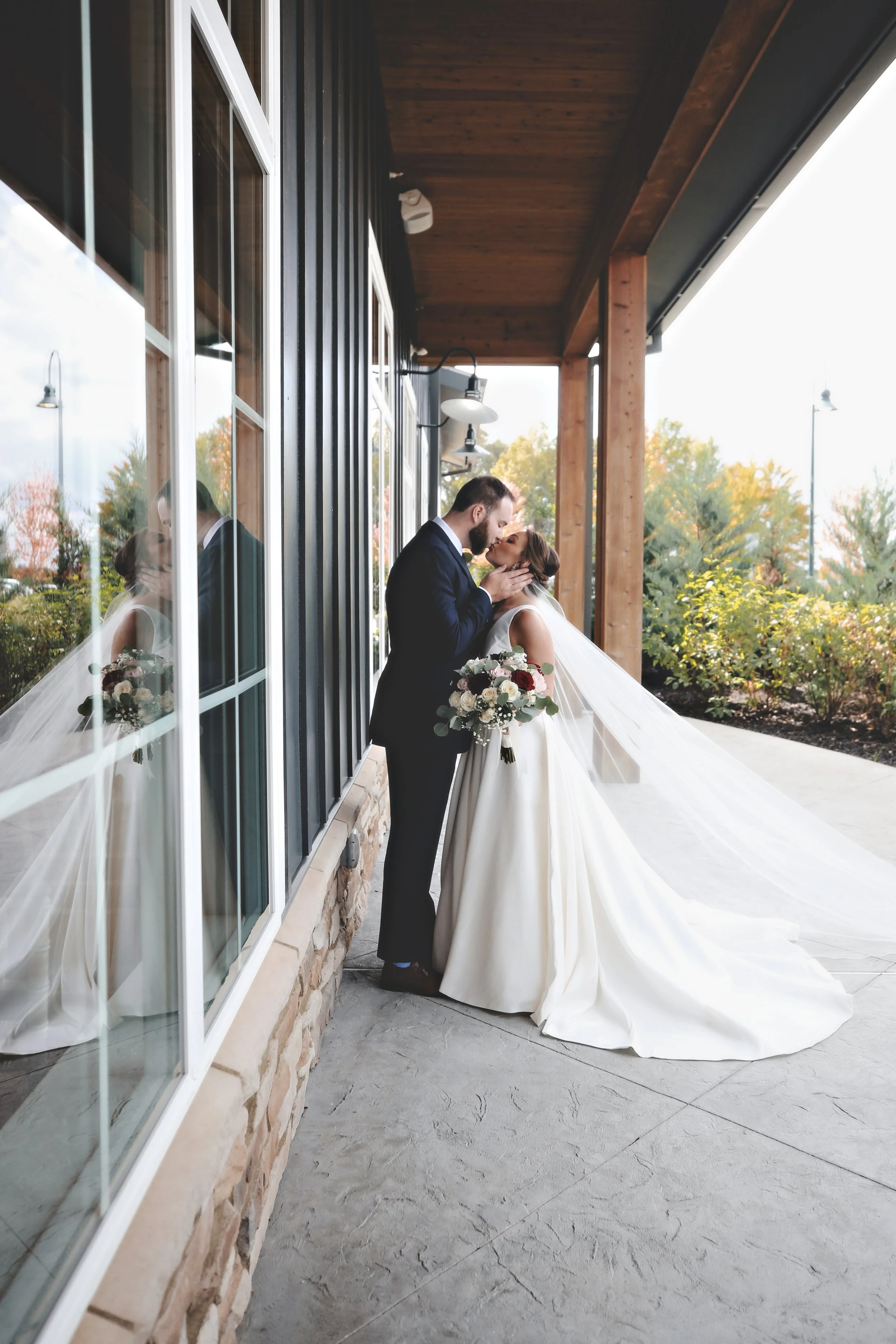 A bride and groom kissing outside a building with large windows and wooden accents, with the bride holding a bouquet of white and red flowers.