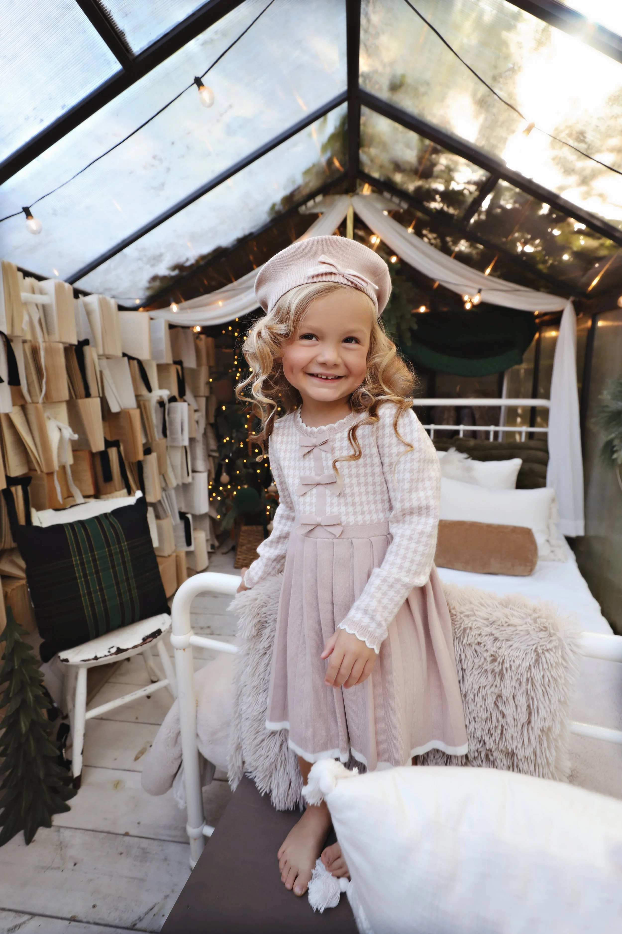 A young girl with long, curly blonde hair wearing a pink beret, a pink dress with bows, and a long-sleeve top, smiling inside a cozy, decorated glass house with fairy lights, cushions, and blankets.