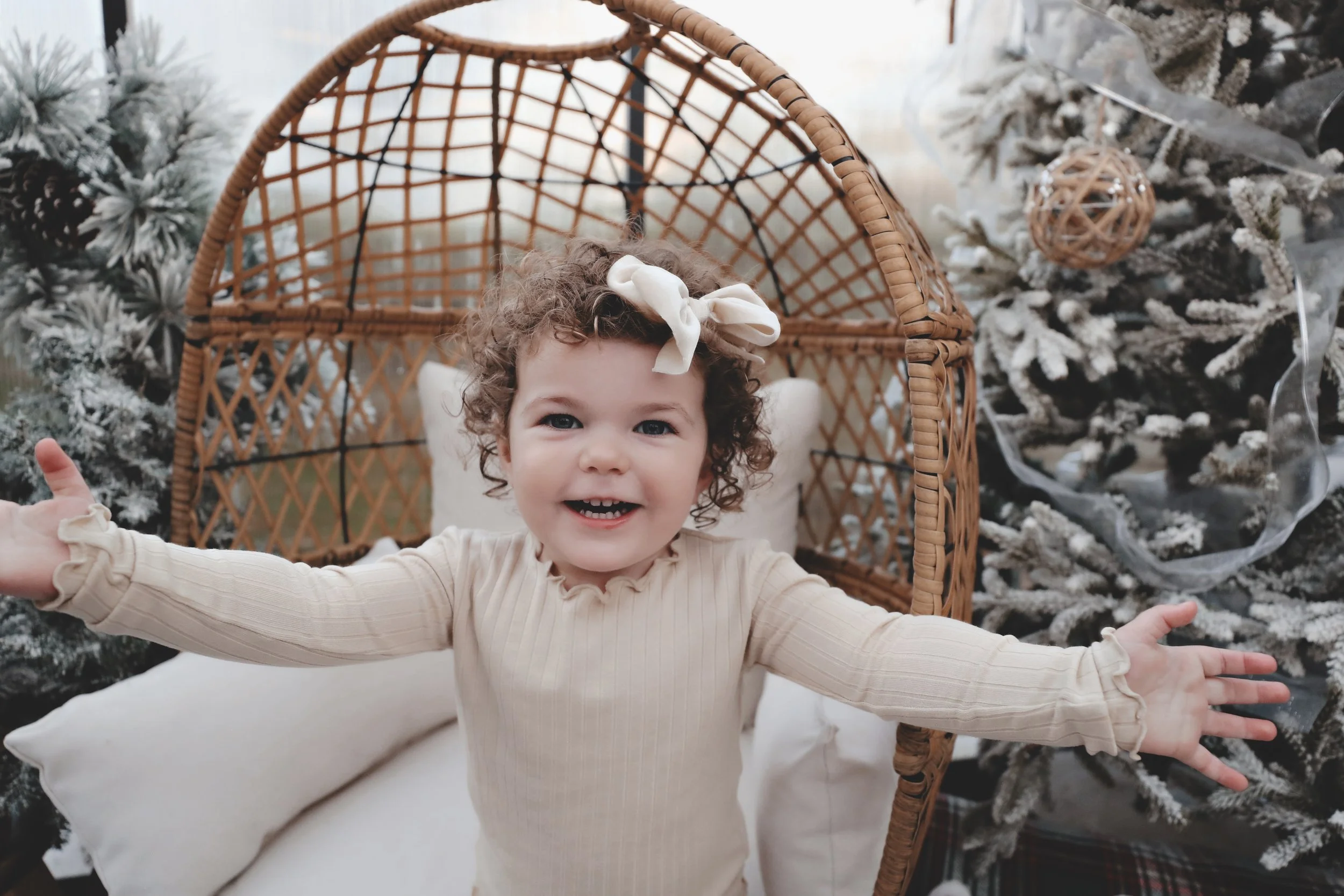 A happy young girl with curly hair and a beige bow is standing with her arms outstretched in front of a christmas tree decorated with snow-like flocking and ornaments, in a cozy indoor setting.