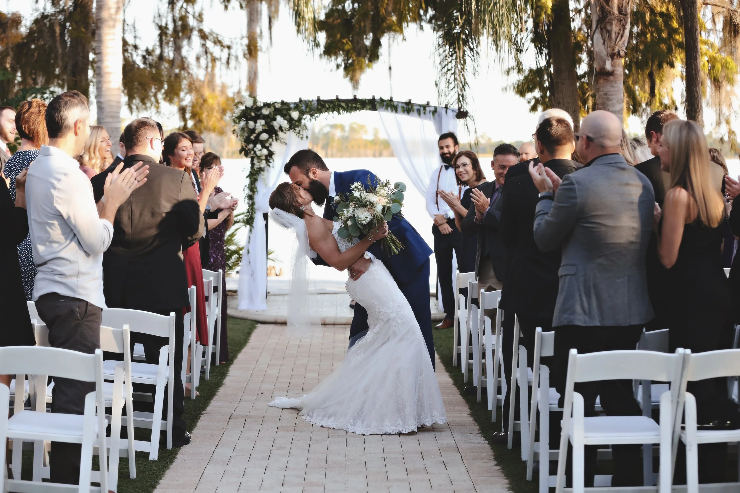 A wedding ceremony outdoors with a bride and groom kissing, surrounded by seated guests clapping, under a decorated arch near a lake, with trees in the background.