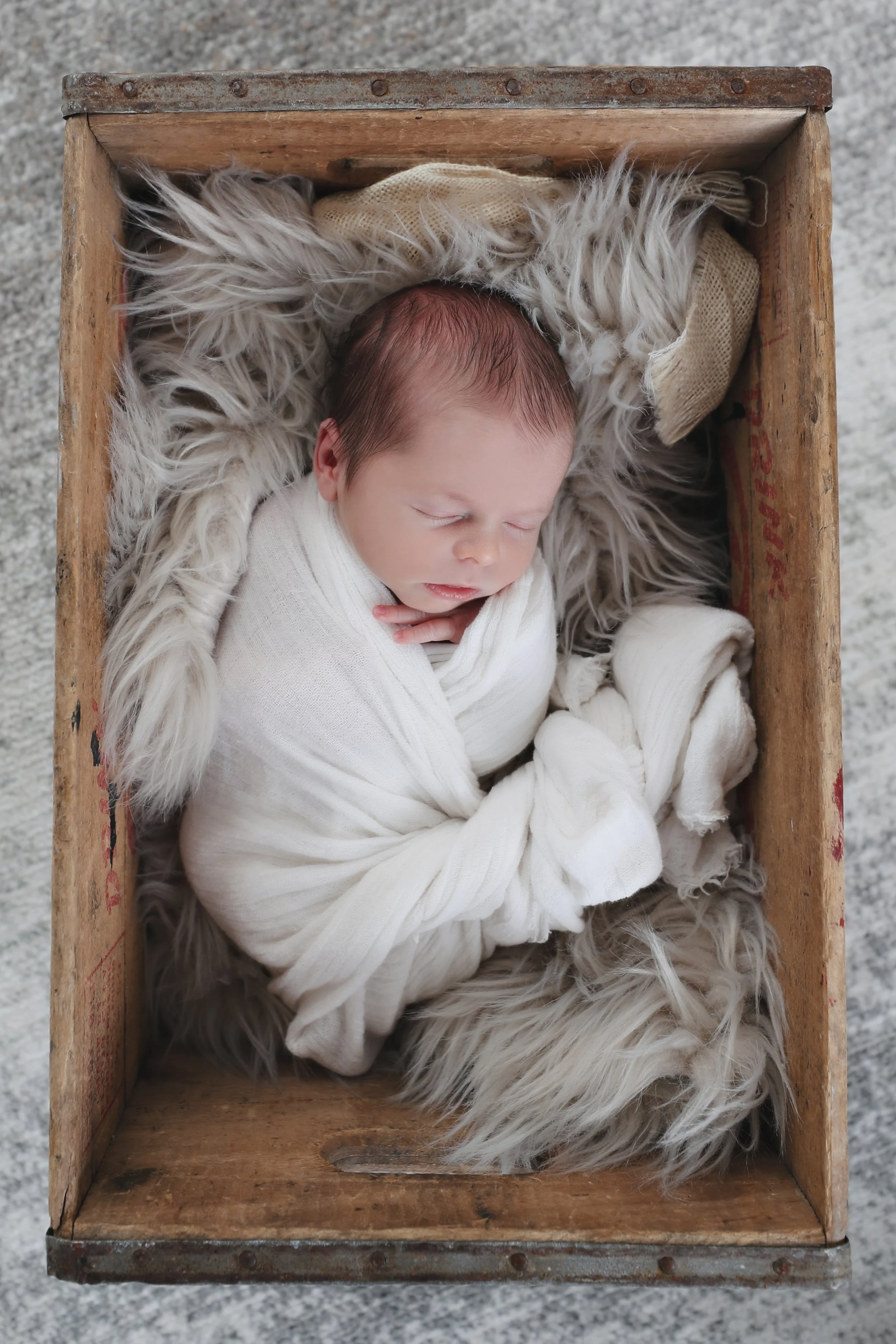 A sleeping baby swaddled in white blankets lying in a wooden crate lined with faux fur and fabric, viewed from above.