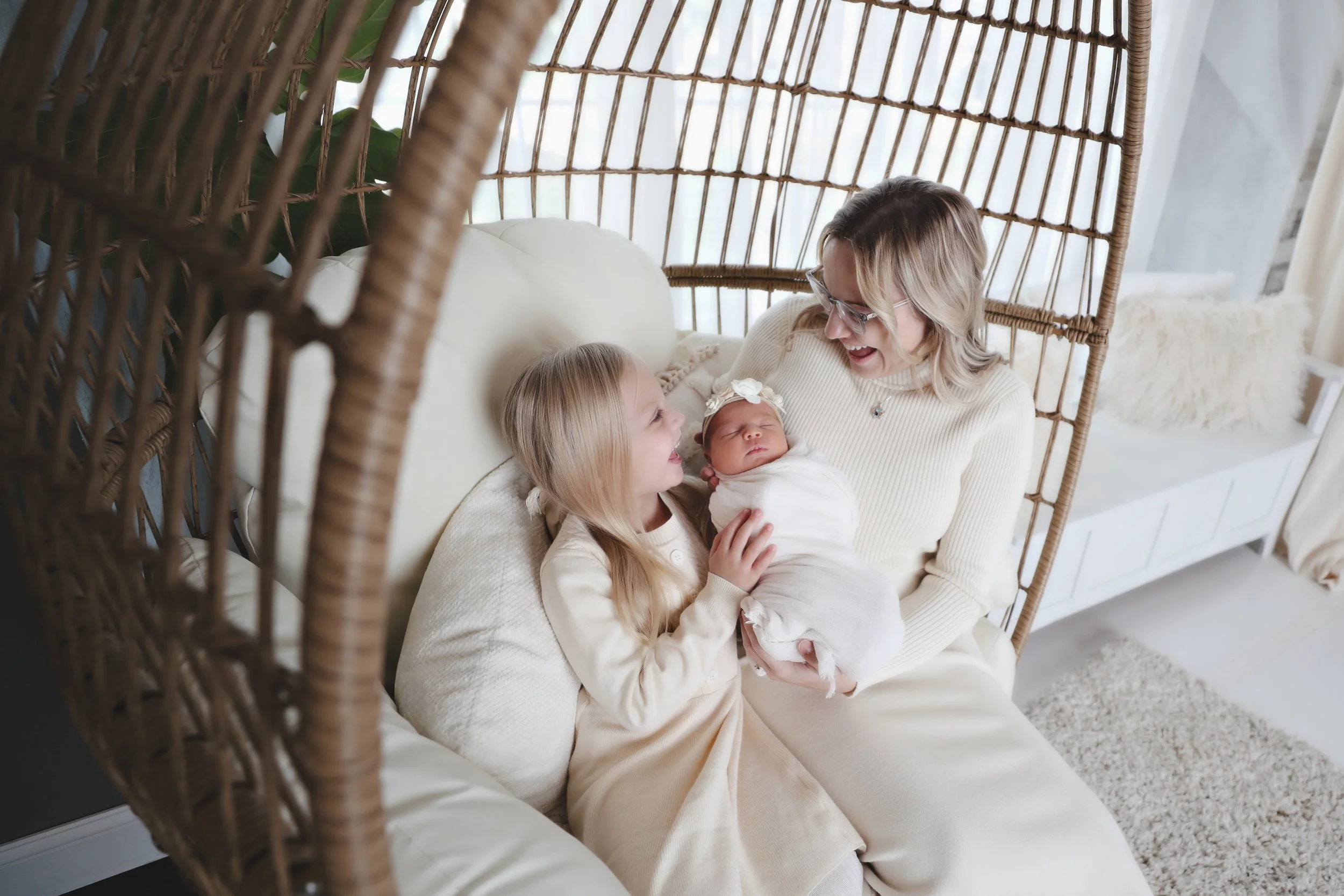 A woman and a young girl holding a newborn baby inside a wicker hanging chair, smiling and looking at each other in a cozy, light-filled room.