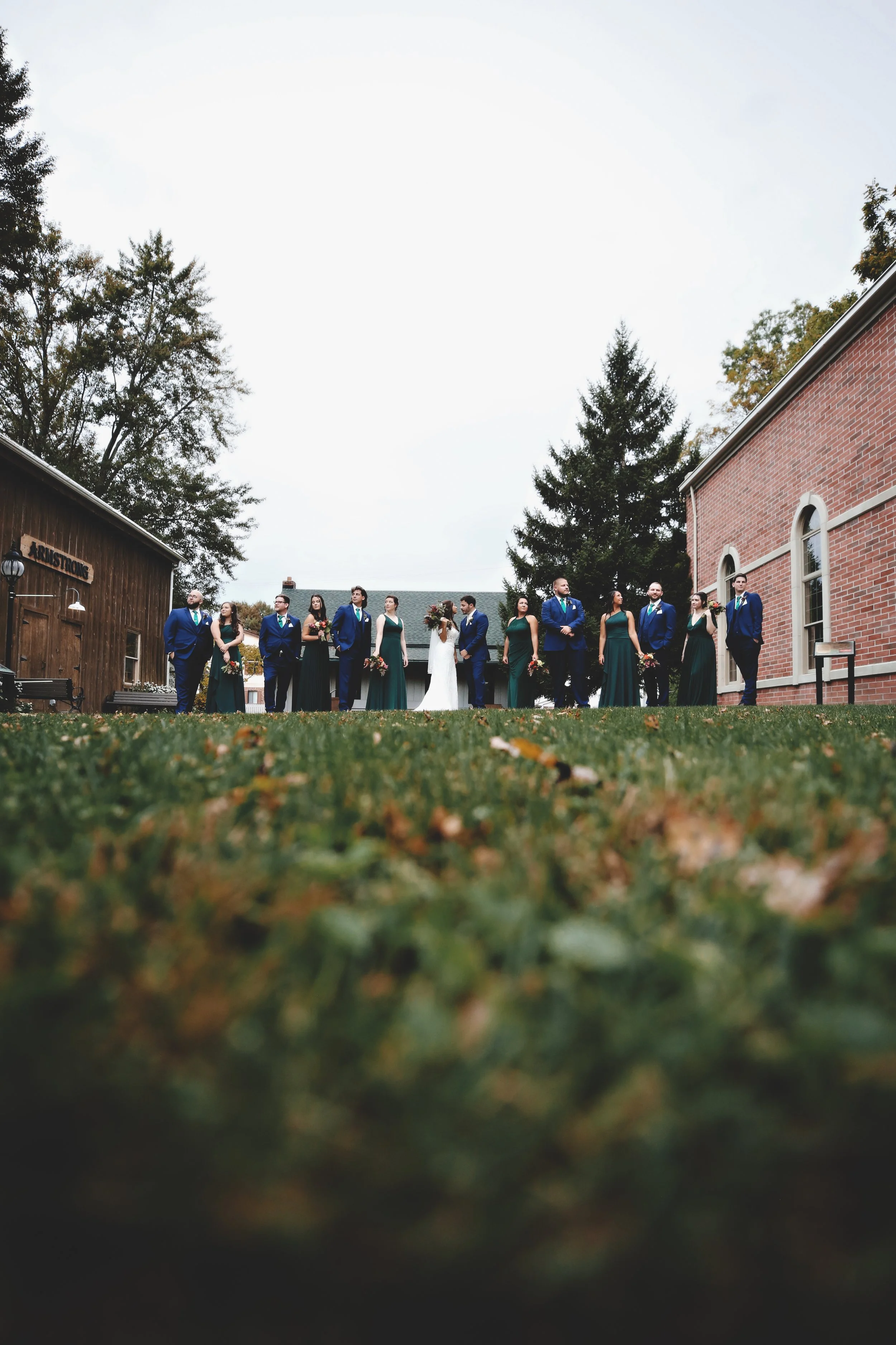 A wedding party posing outdoors in front of a building with guests and trees in the background.