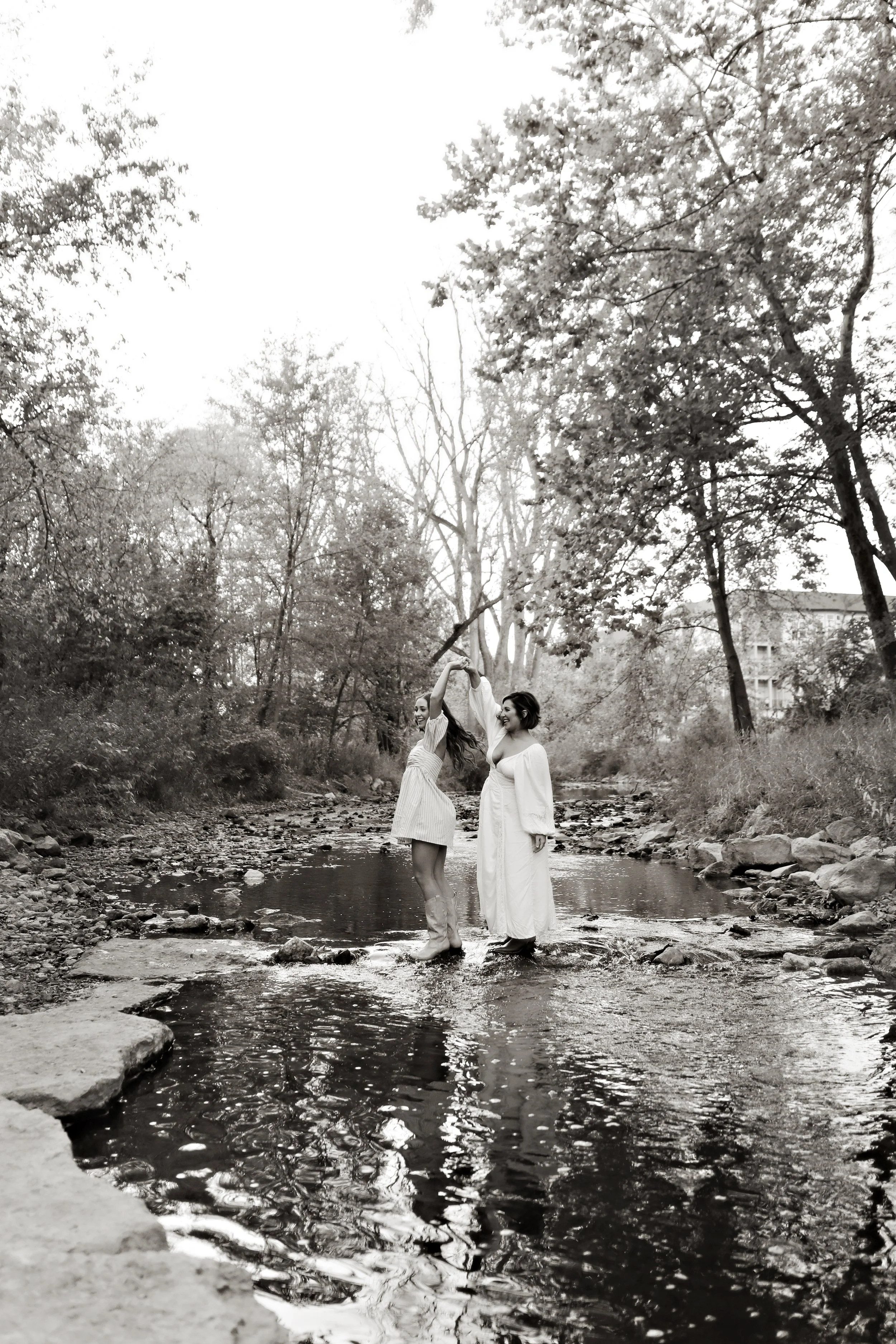 Two women standing in a shallow creek surrounded by trees, holding hands and smiling, in a black and white photo.