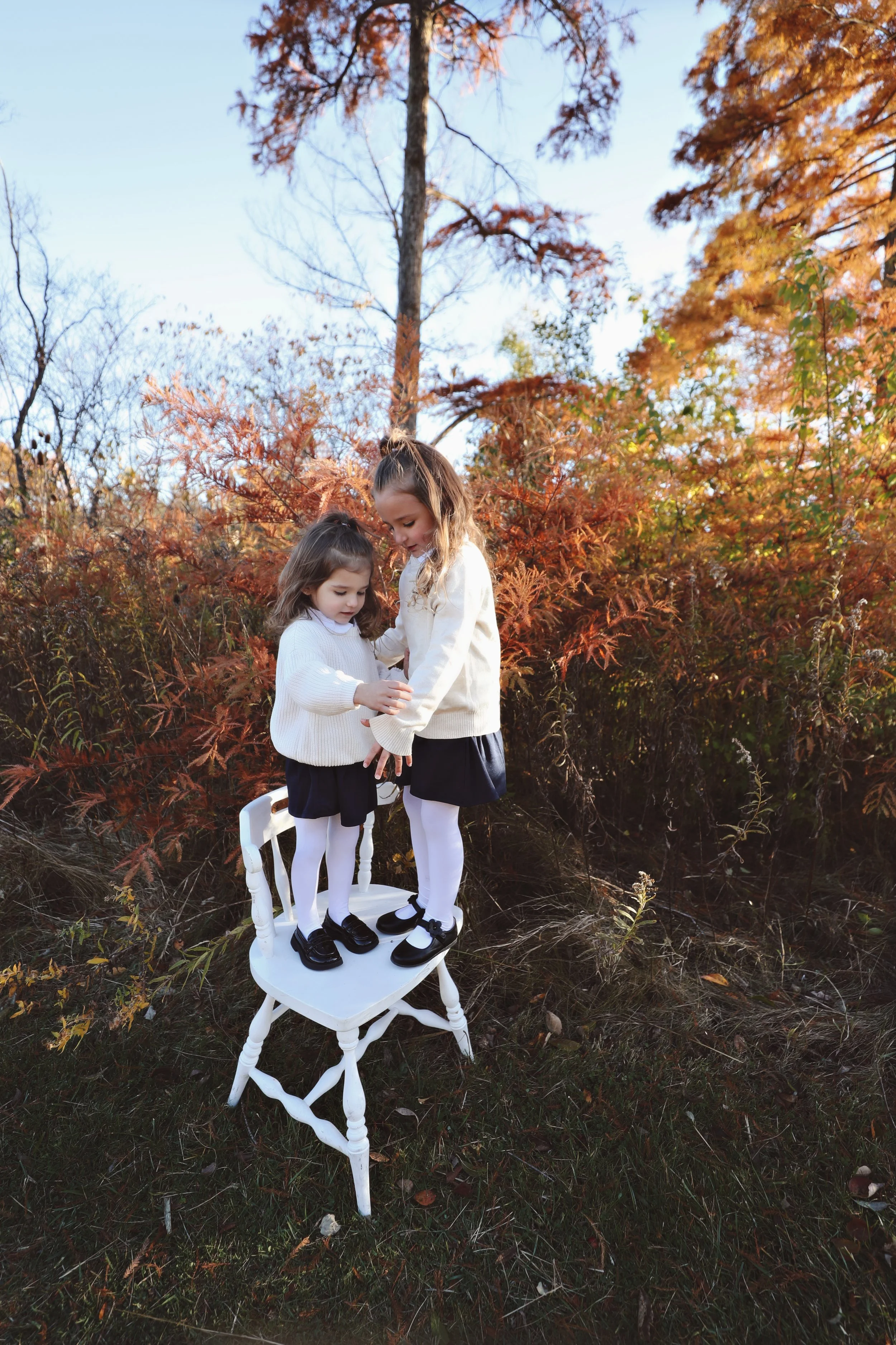 Two young girls standing on a white chair outdoors in a fall setting, wearing white sweaters, navy skirts, white tights, and black shoes, surrounded by orange and yellow autumn leaves.