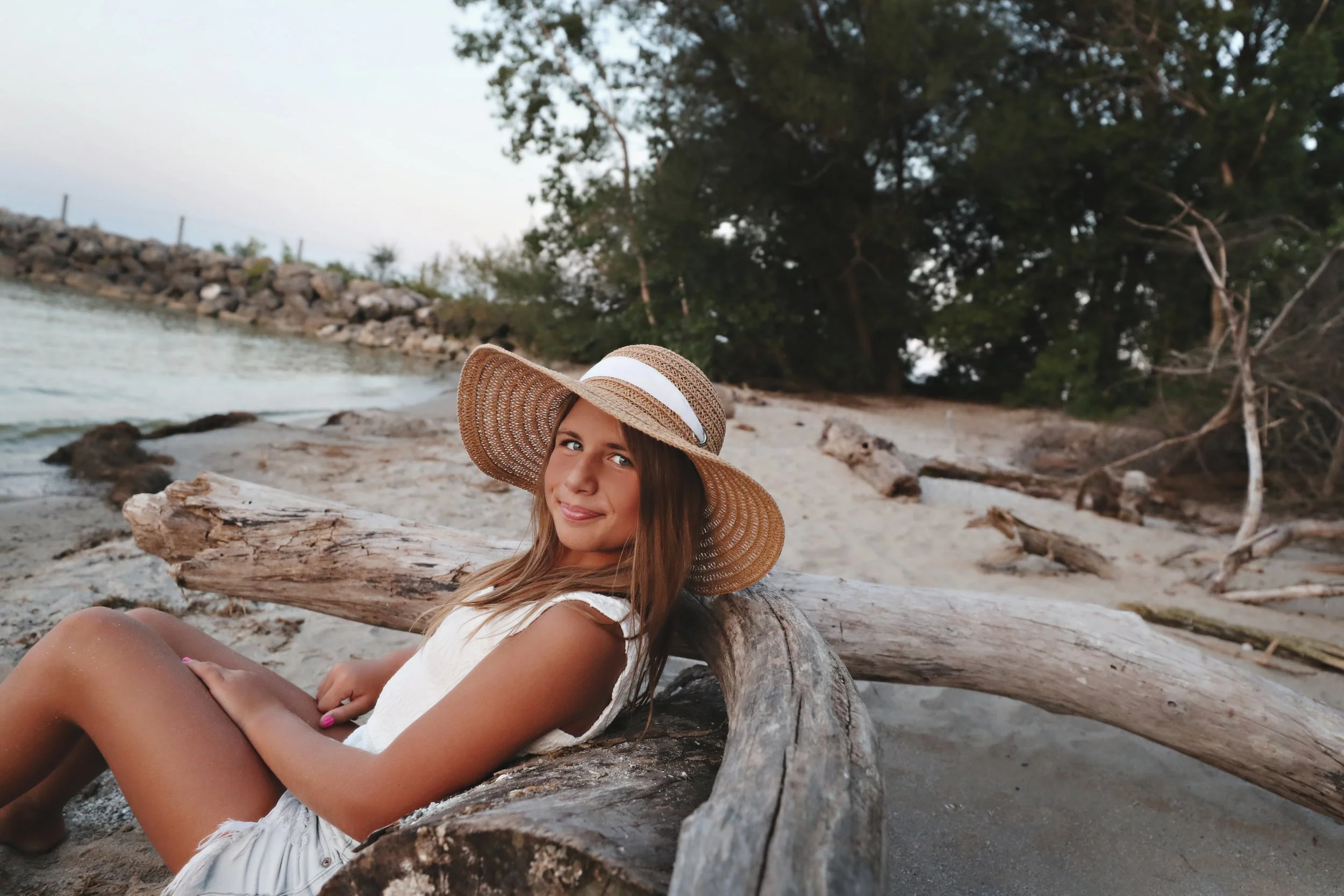 A young girl wearing a wide-brimmed hat sitting on a large piece of driftwood on a beach with sand, trees in the background, and water nearby.