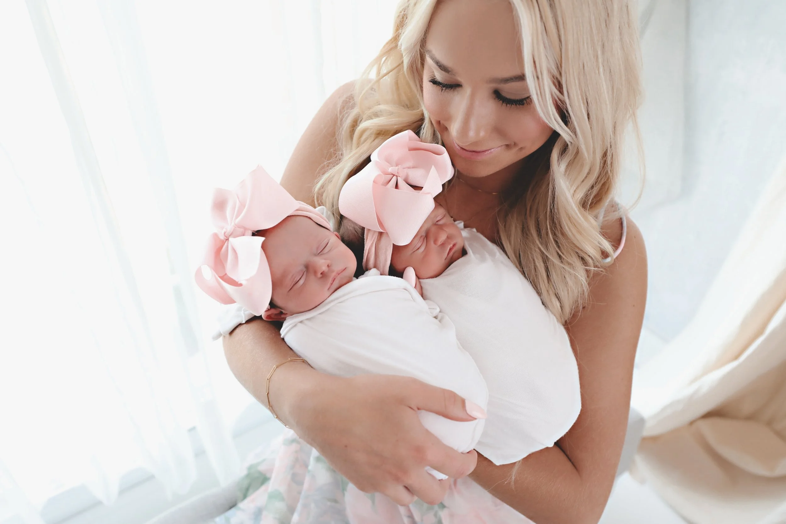 A woman holding two newborn babies wrapped in white blankets, each wearing a pink bow headband, near a window with sheer curtains.
