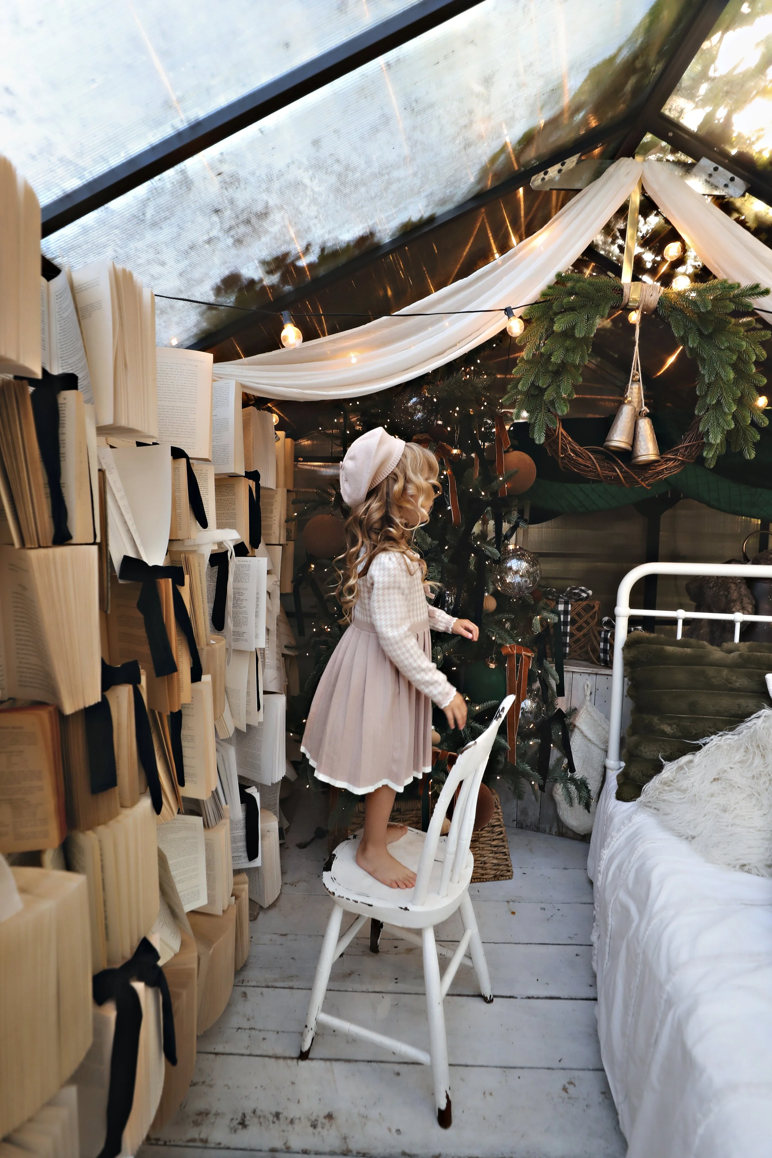 A young girl with curly blonde hair, wearing a beige dress and white beret, standing on a white wooden chair inside a cozy, decorated room with a Christmas tree, books, and festive decorations.