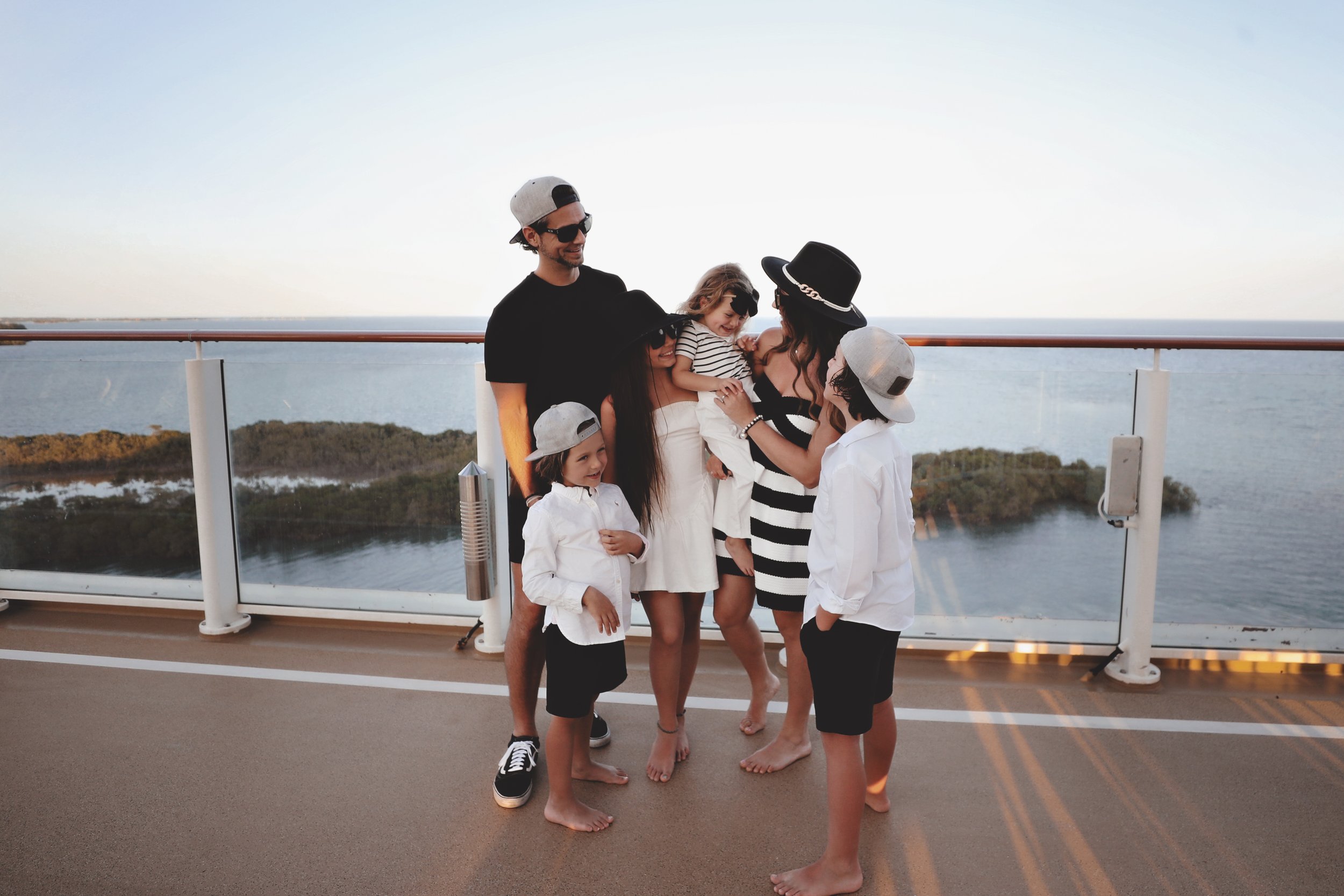 A family of six with three children and three adults in casual summertime clothing, standing barefoot on the deck of a ship with a scenic ocean view in the background. The children are wearing caps and the adults are dressed in black and white striped and solid clothing, with some wearing hats and sunglasses. The family appears to be enjoying a moment together, smiling and talking.
