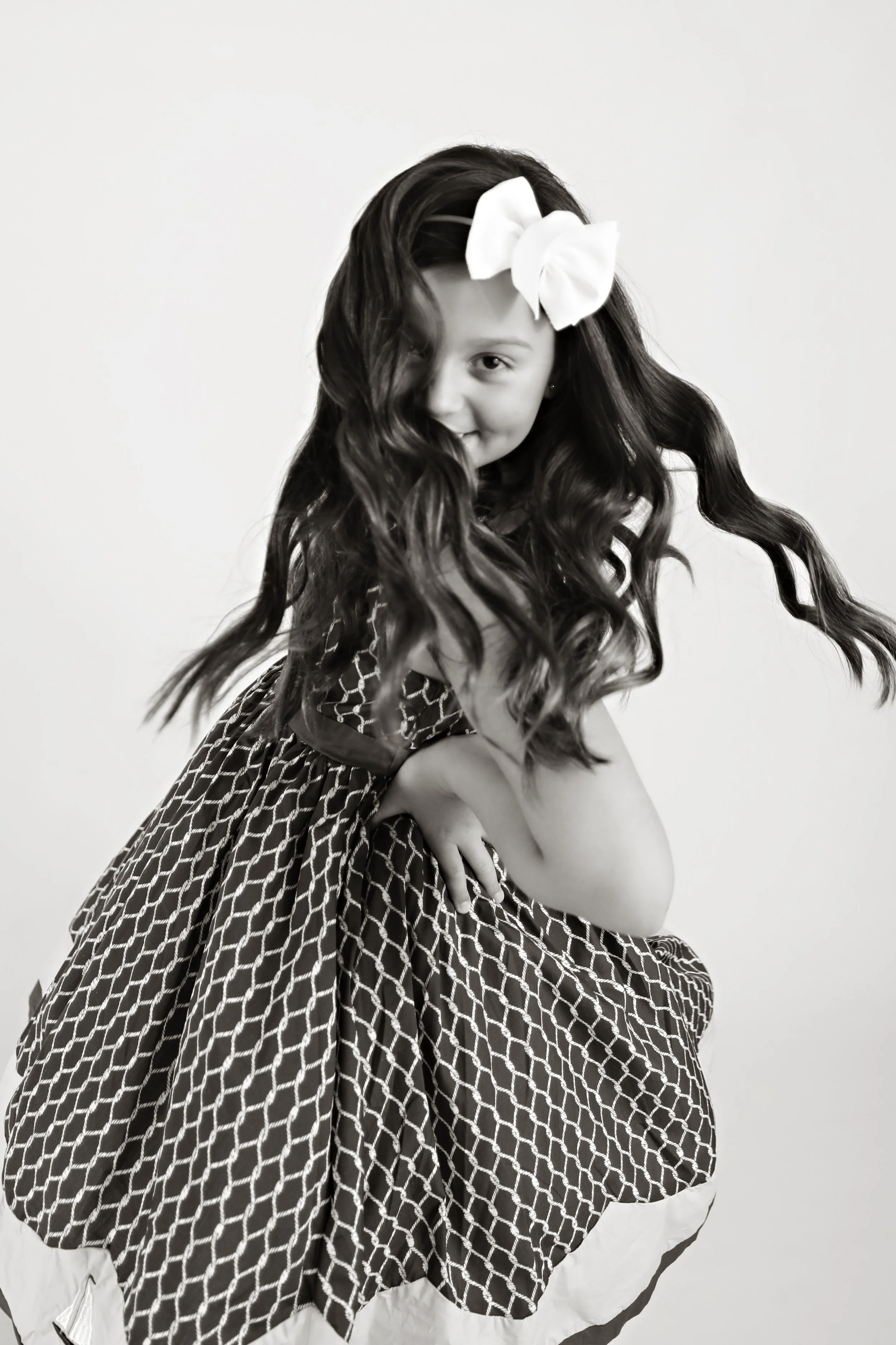 A young girl with long wavy hair, wearing a large bow on her head, posing and smiling in a black-and-white photo.