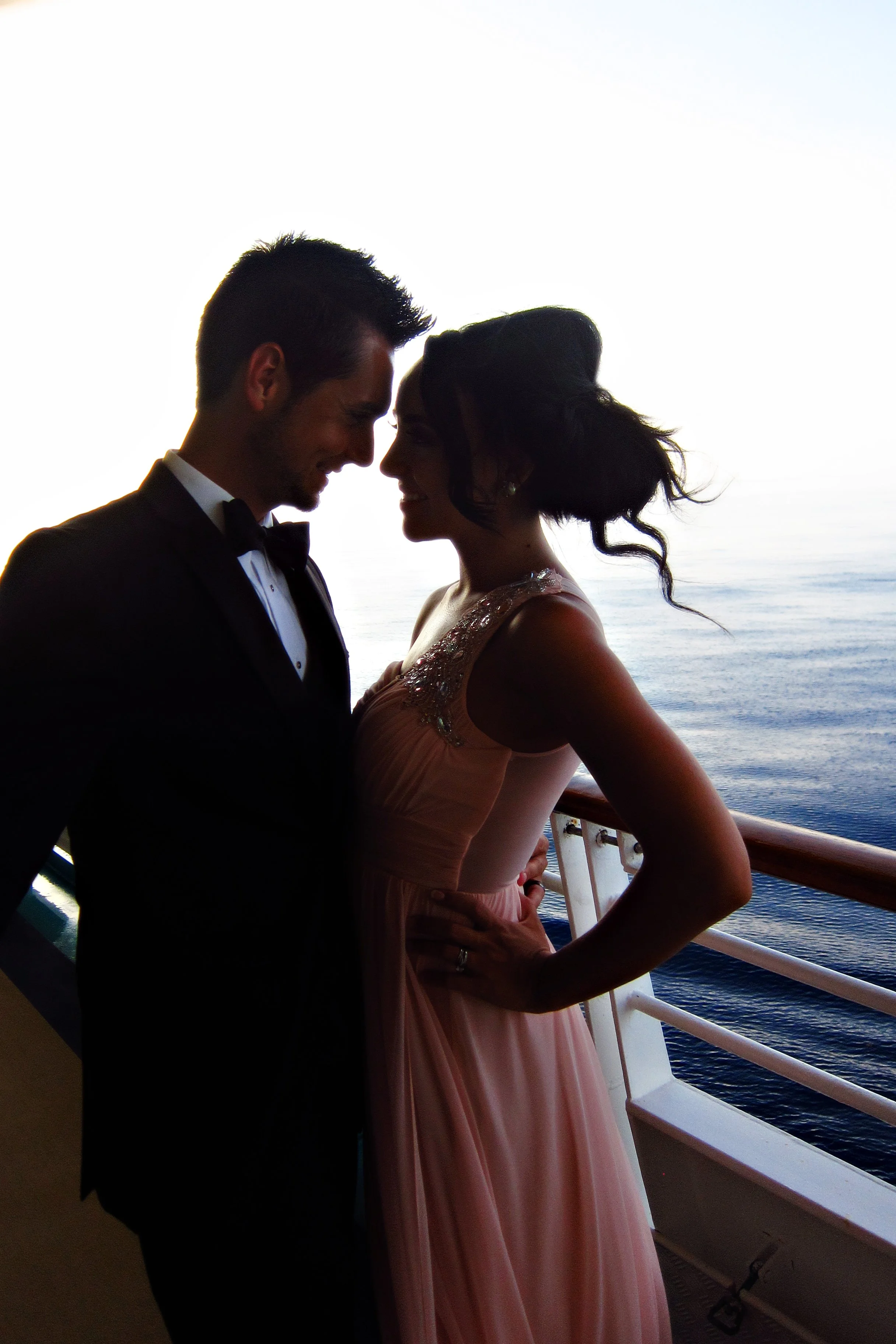 A couple dressed in formal attire on a boat, embracing with their foreheads touching, against the backdrop of the sea and sky.