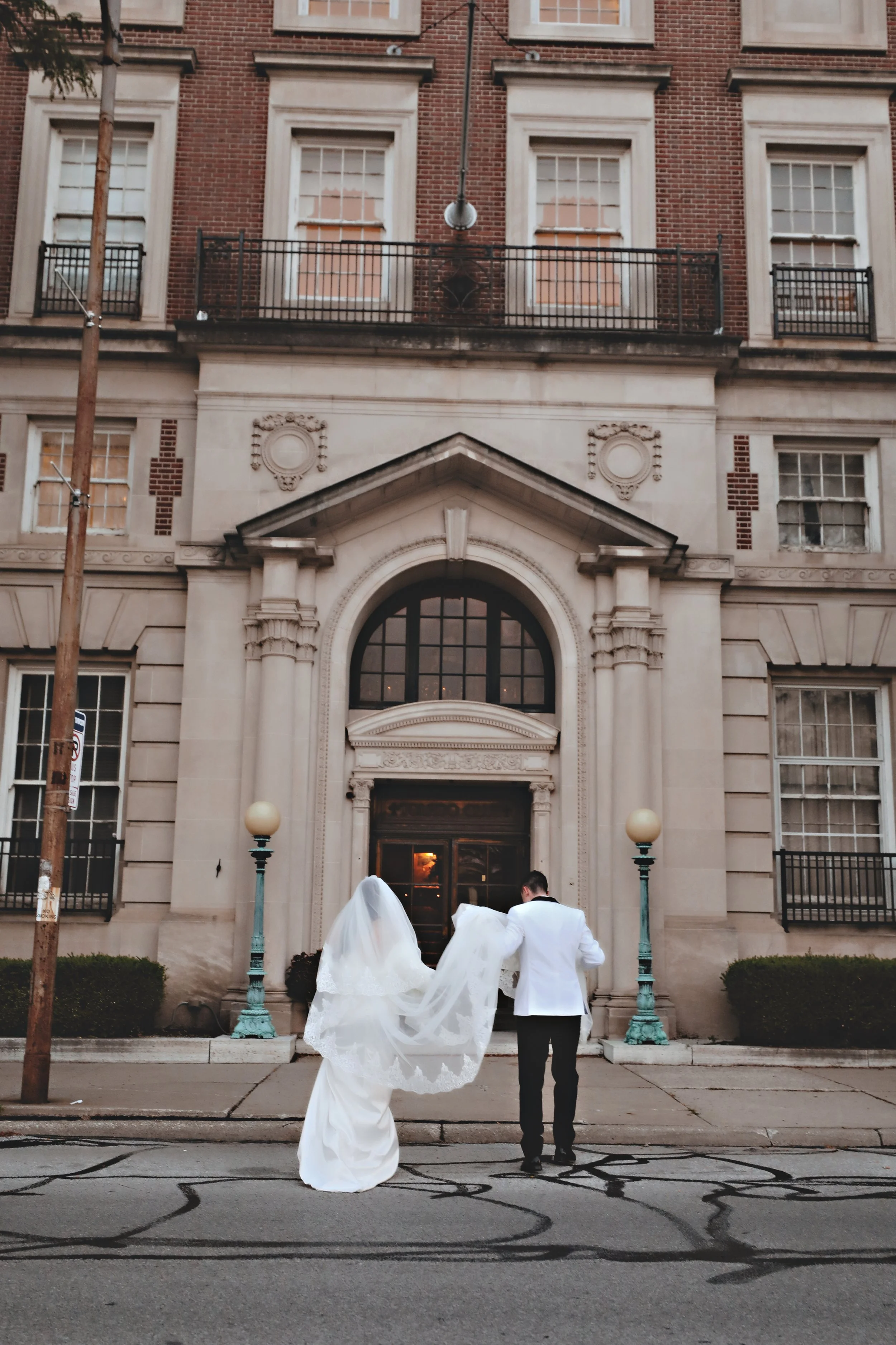 A bride in a white wedding gown and veil, and a groom in a white tuxedo, standing in front of a historic brick building with large windows and ornate stone details.
