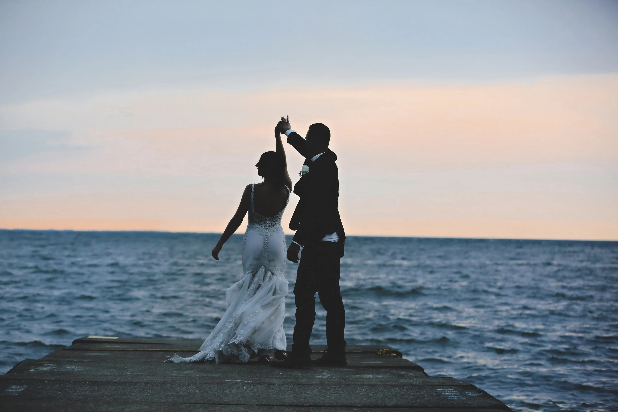 Silhouette of a bride and groom dancing on a wooden pier by the sea during sunset.