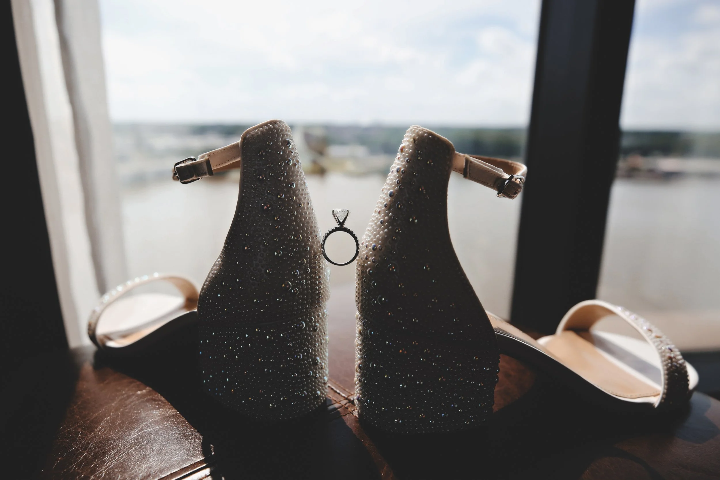 A pair of beige high-heeled shoes decorated with small crystals, with a diamond engagement ring hanging between the heels, set against a window overlooking a large body of water.