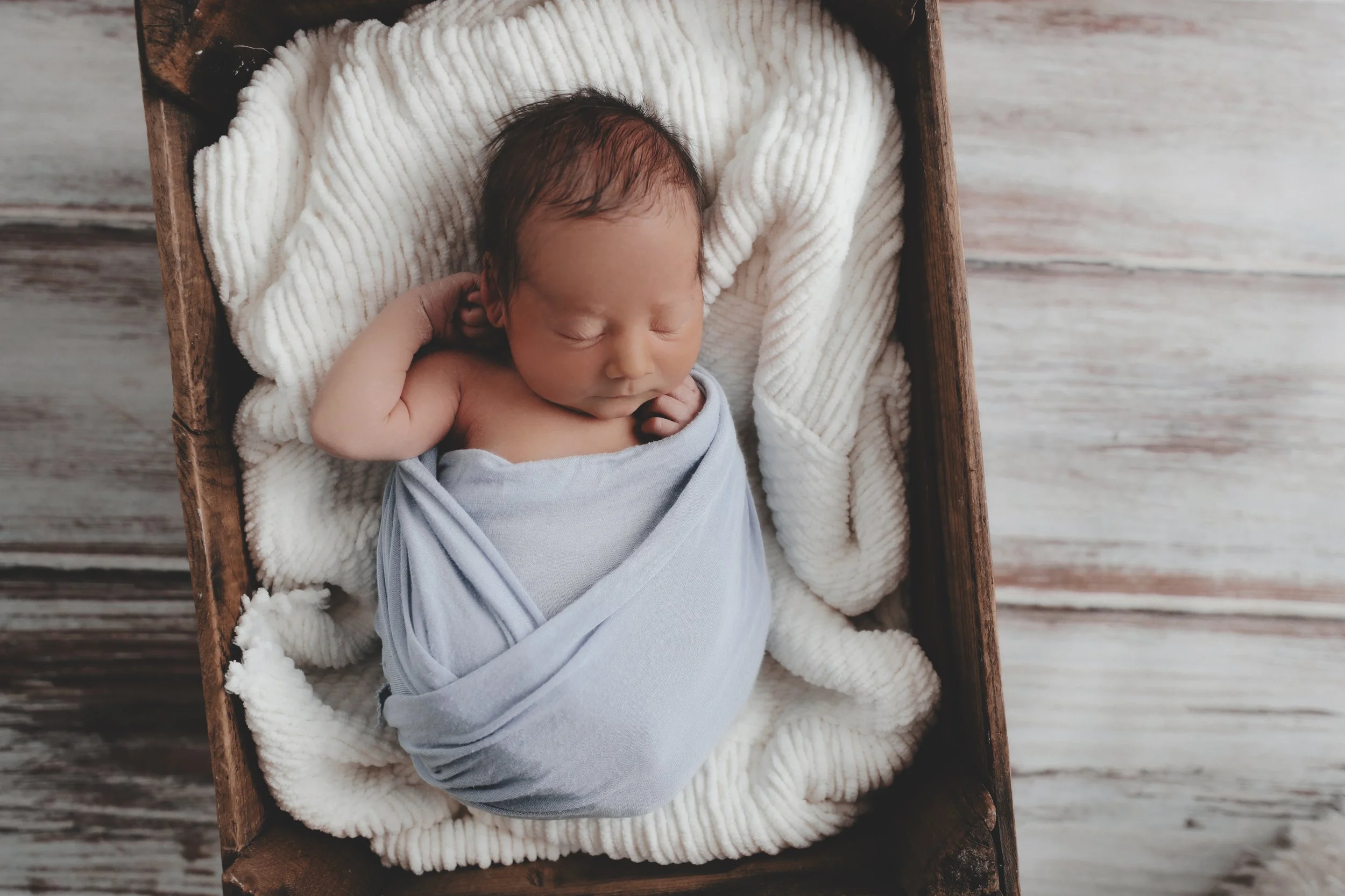 A sleeping newborn baby swaddled in a light blue blanket, lying in a wooden basket lined with a white, textured blanket, on a wooden floor.