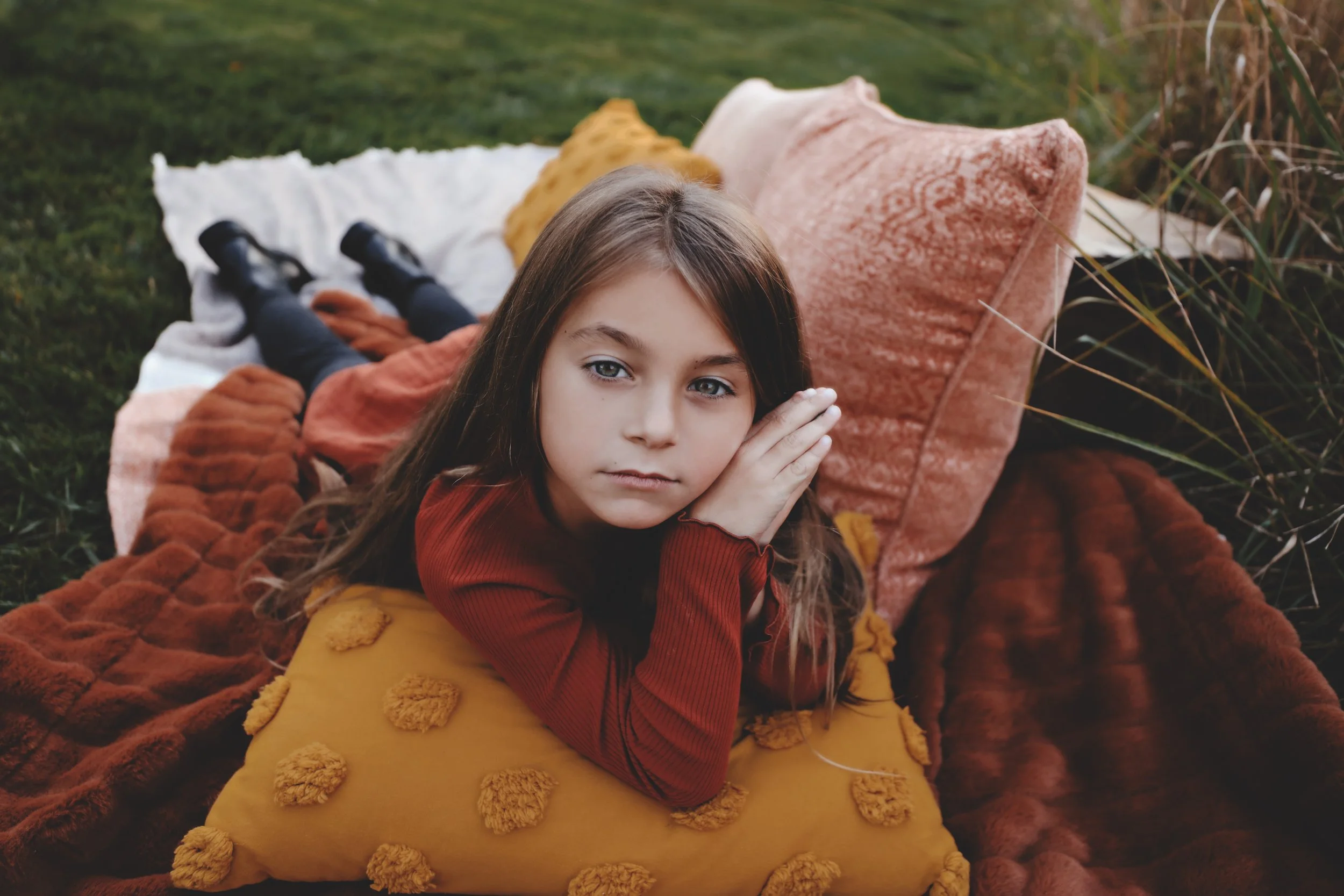 Young girl lying on her stomach on a blanket outdoors, resting her face on her hand, surrounded by colorful cushions.