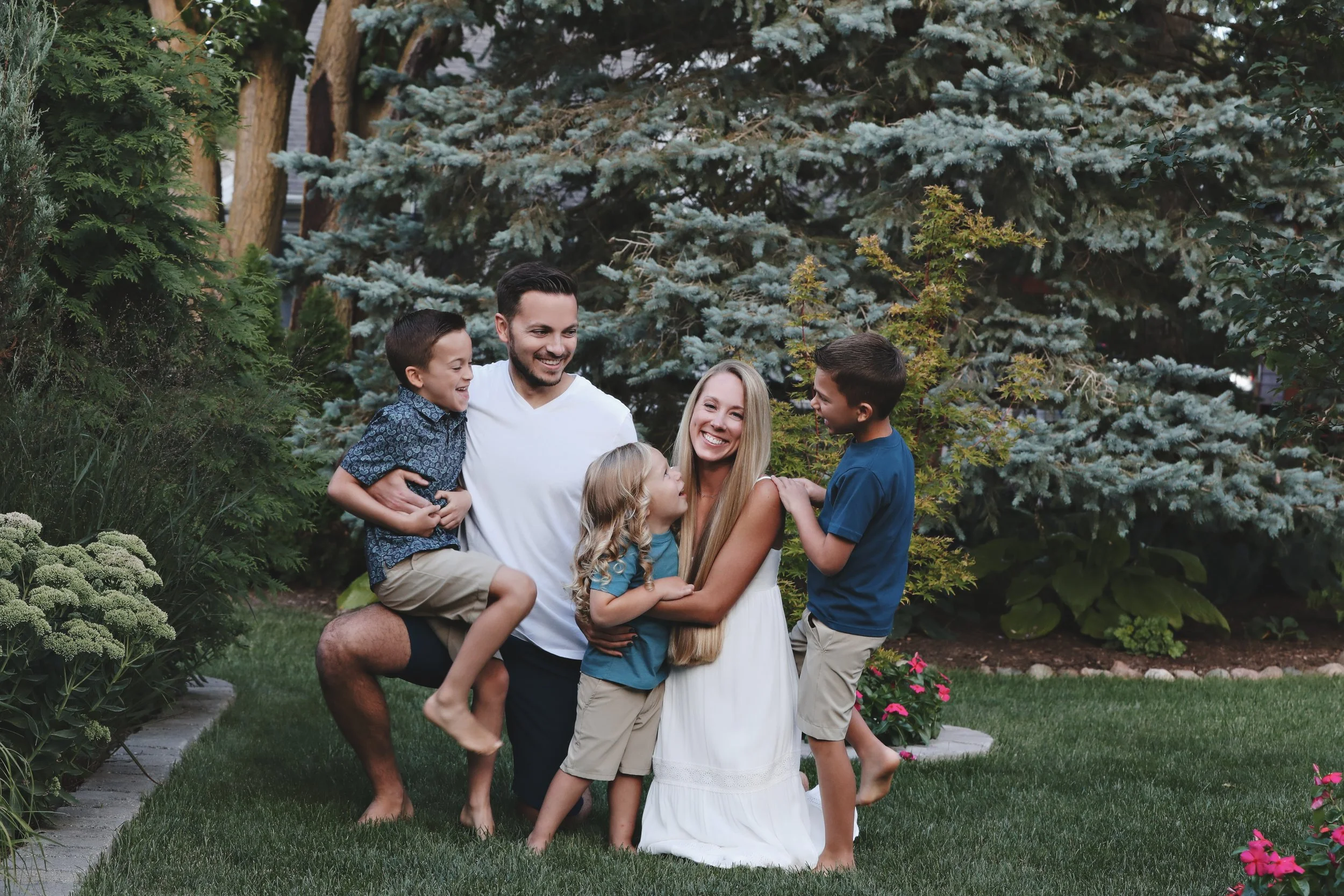 A family of five, including a father, mother, and three children, is outside in a garden with lush greenery and trees. They are smiling, embracing, and playing together on the grass.