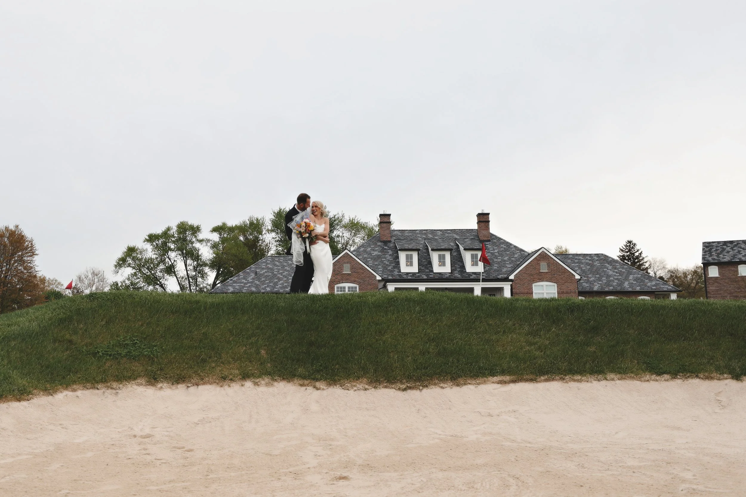 A newlywed couple standing on a grassy area near a sand trap on a golf course, with a large brick house in the background.