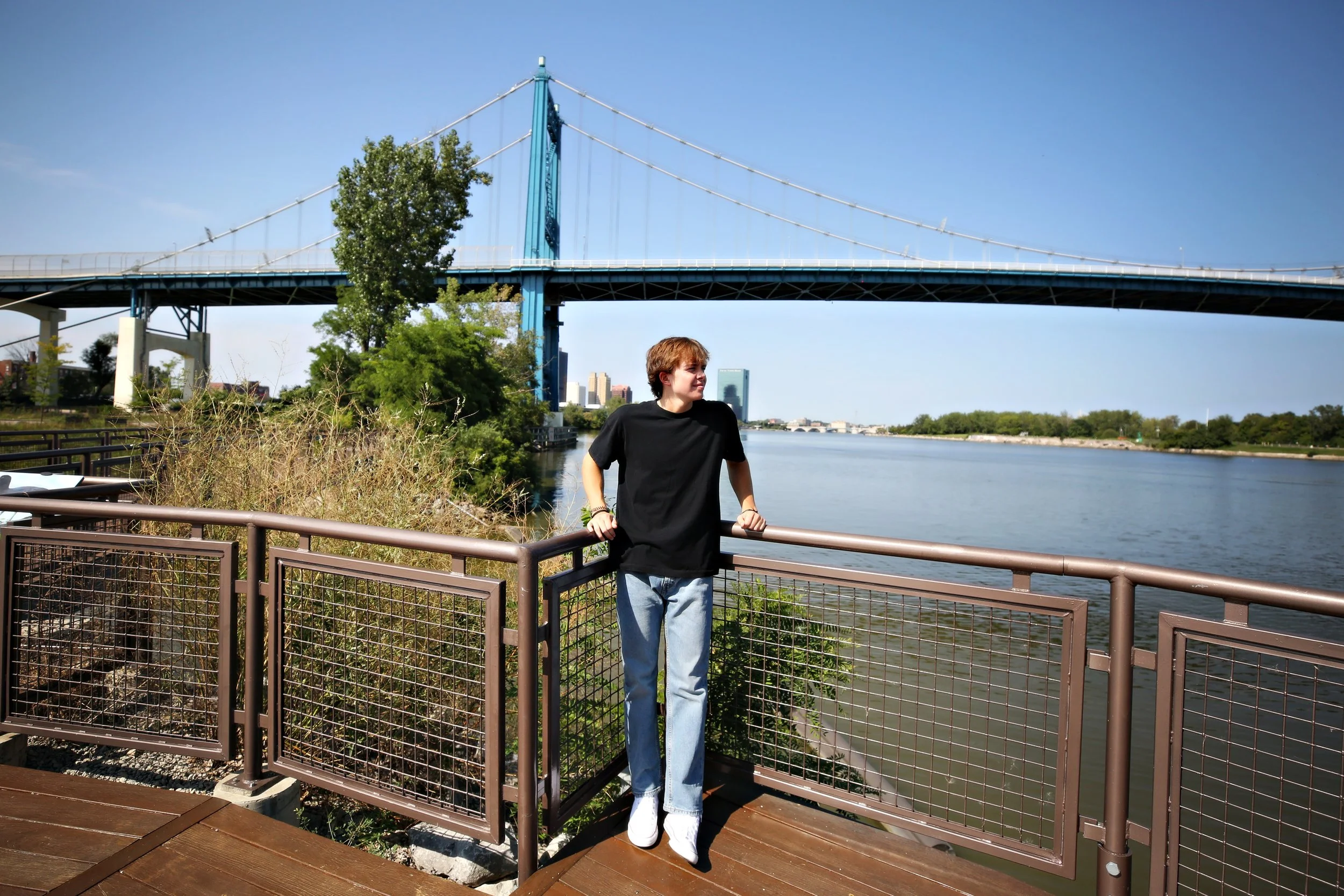 A person in a black t-shirt and light blue jeans standing on a wooden deck by a river with a bridge overhead, greenery, and city buildings in the background.
