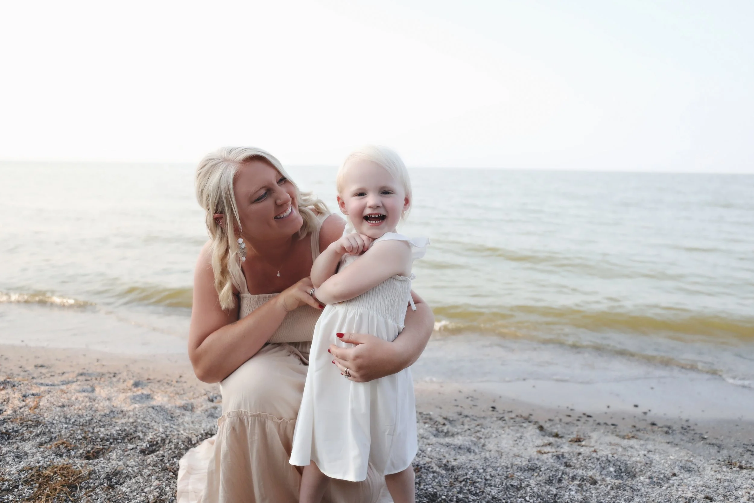 A woman and a young girl laughing and playing together on a beach near the water.